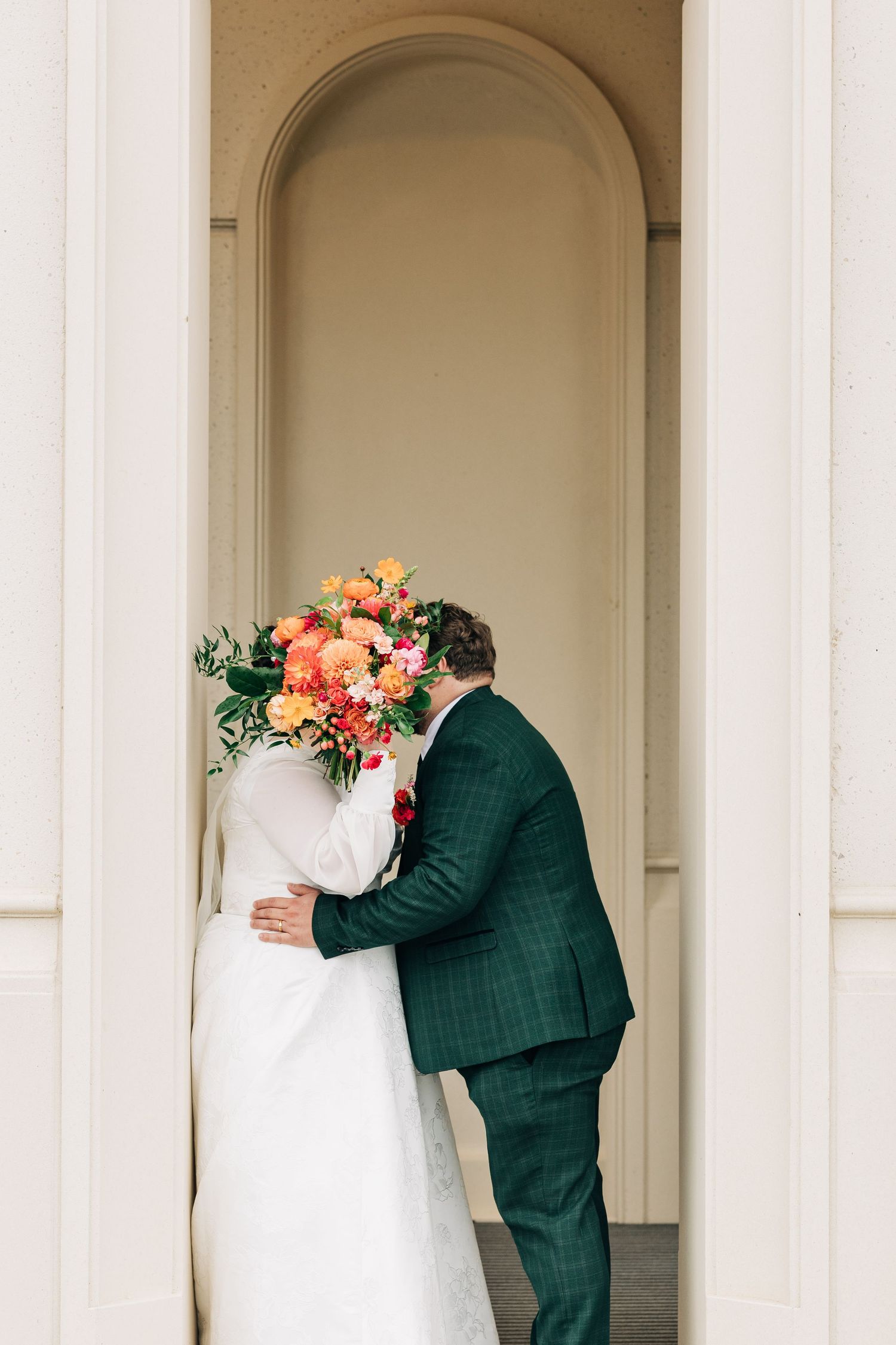 A romantic wedding moment with a green suited figure and white dressed figure embracing in an elegant archway.