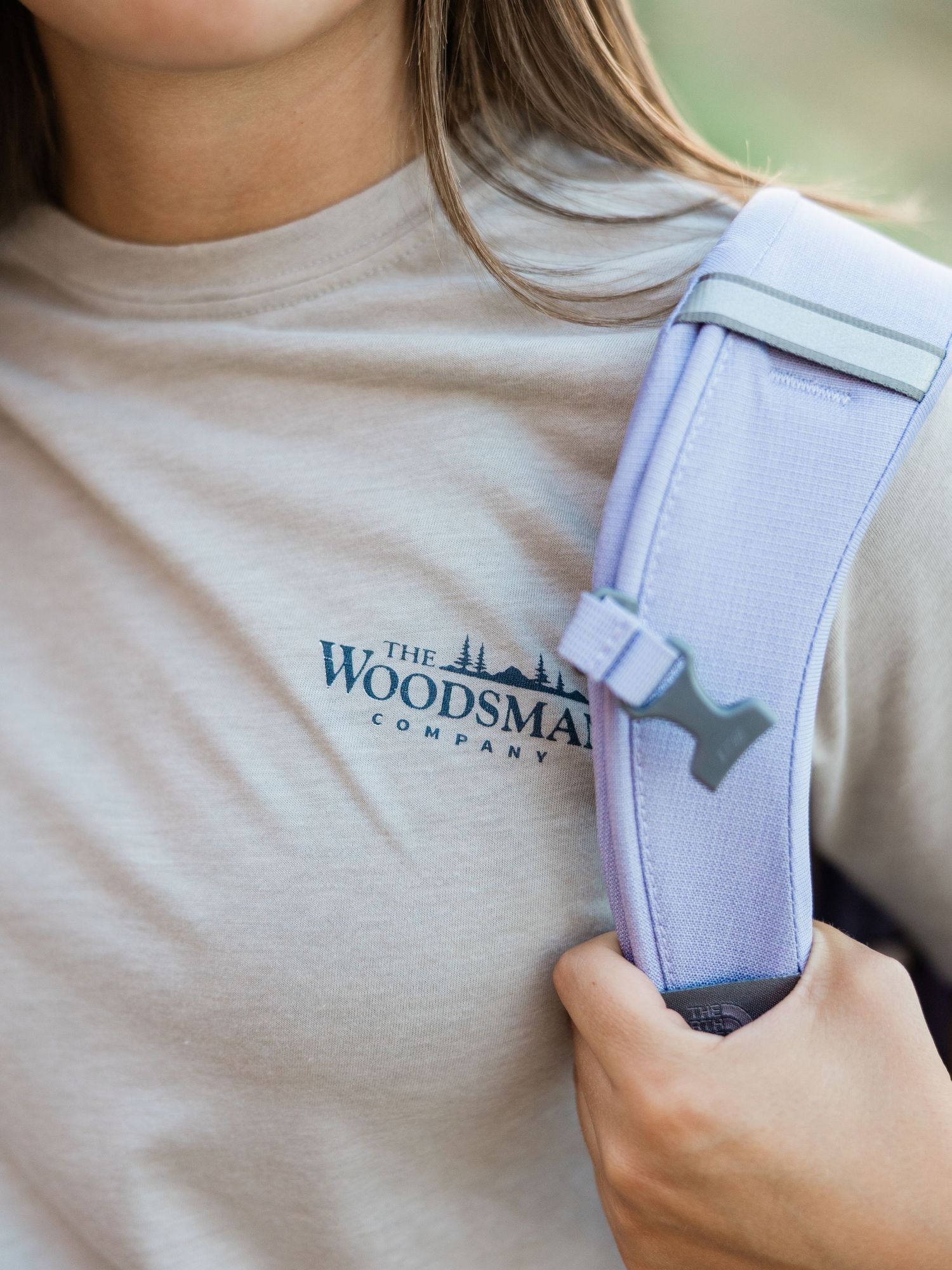 Woman wearing a beige The Woodsman Company t-shirt while holding a lavender backpack strap outdoors.