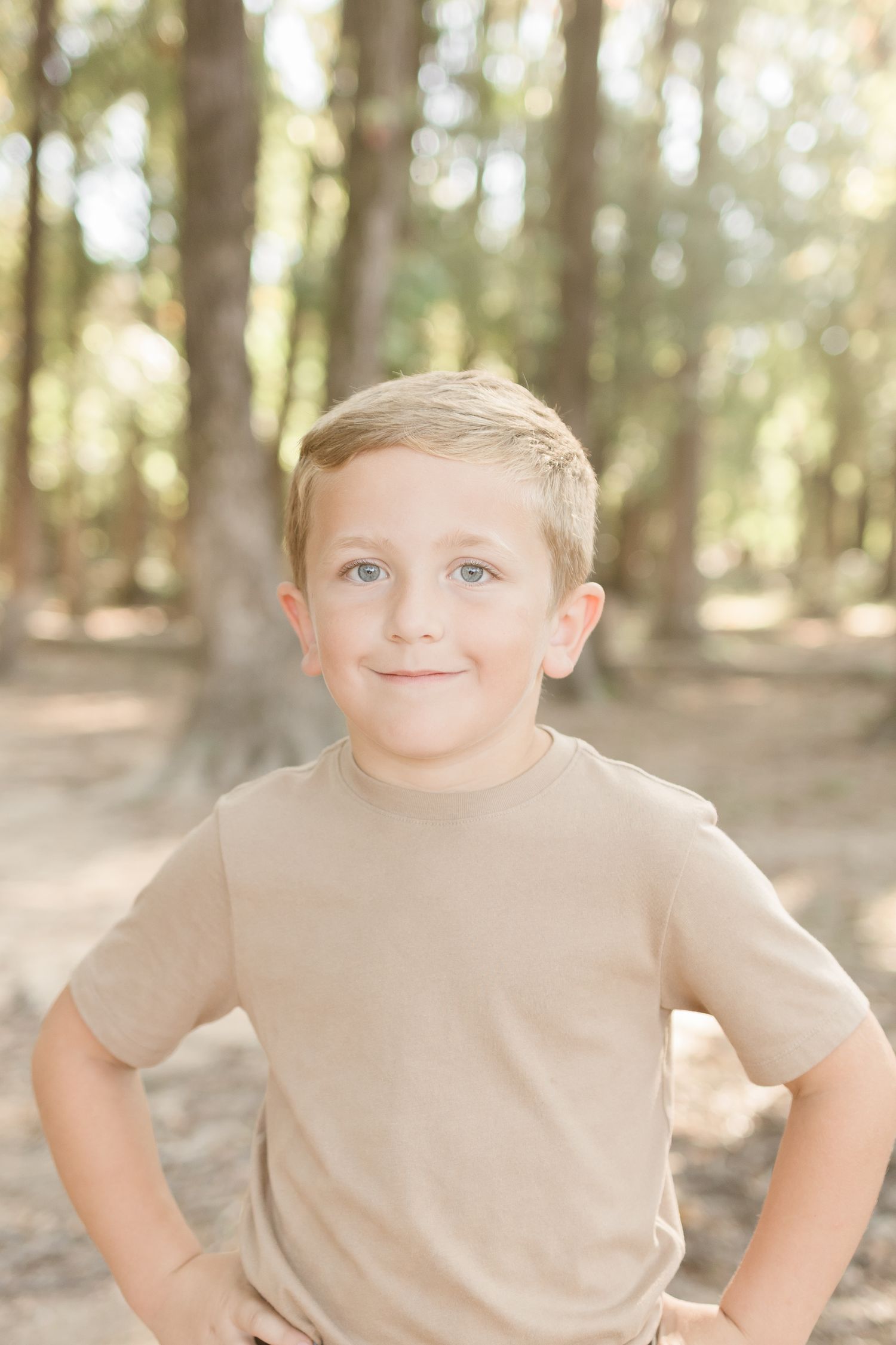 A young child in a beige t-shirt makes silly faces while posing in a sunlit wooded area.