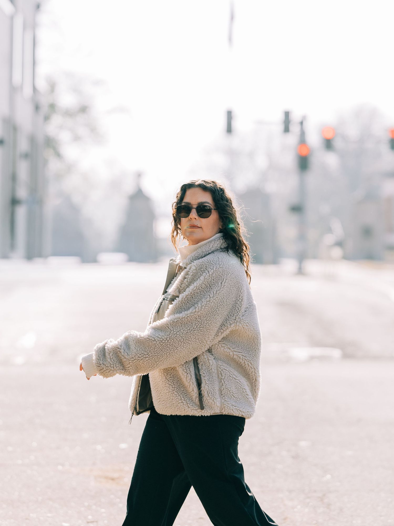 A woman in a cream teddy jacket and black pants crosses a sunlit street, wearing sunglasses, with a blurred urban backdrop.