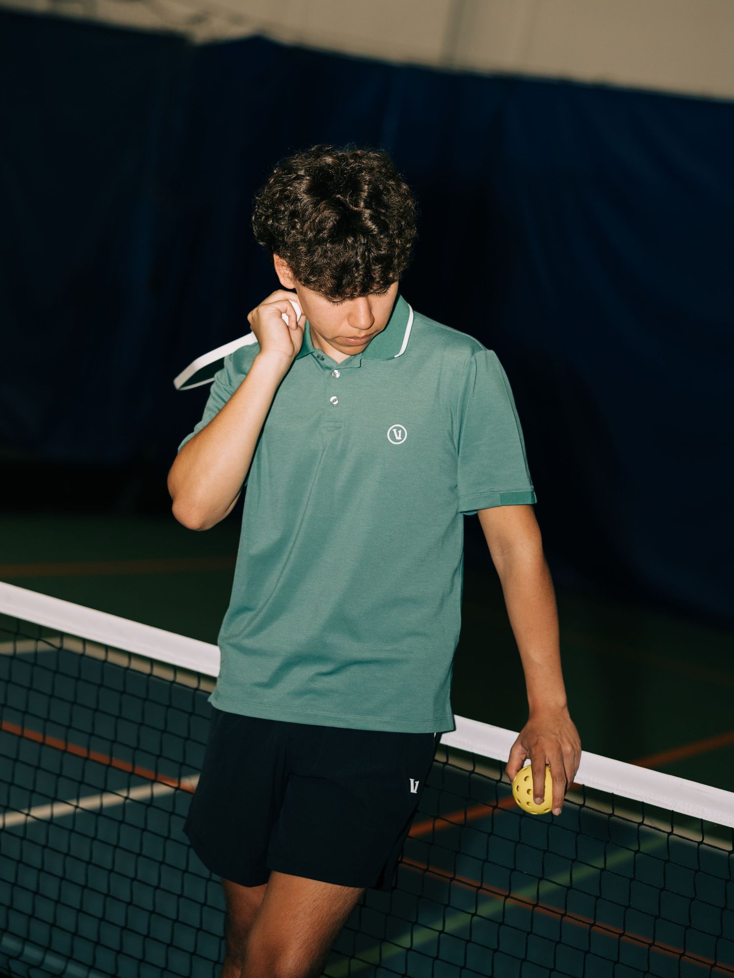 Young man in green polo shirt holding tennis racket and ball near net on indoor tennis court.