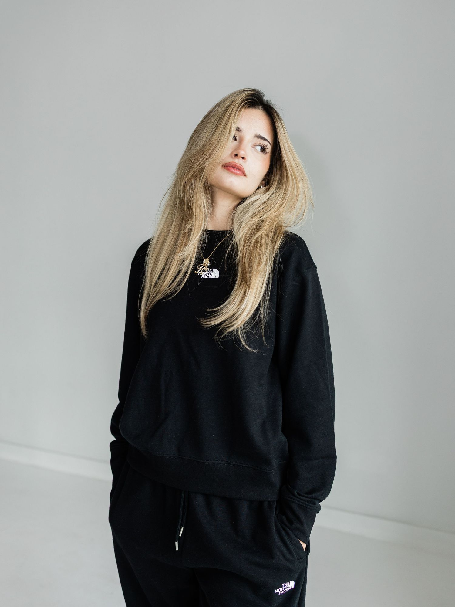 Woman with blonde hair wearing a black North Face sweatsuit poses against a light gray background.