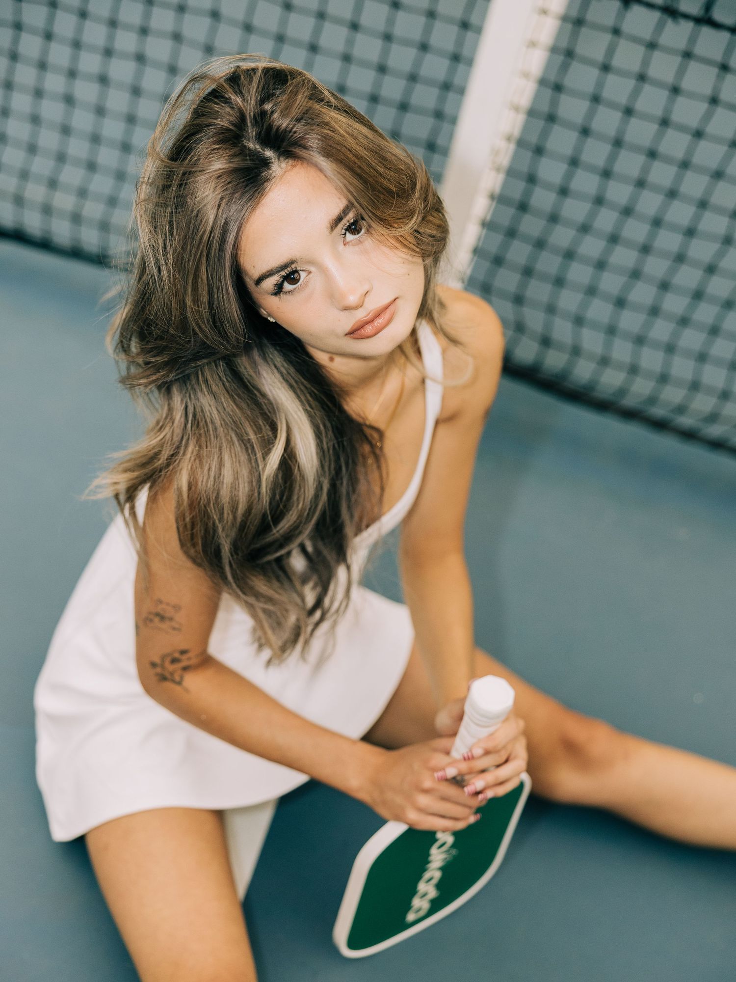 Young woman in white tennis dress sitting on court holding a green pickleball paddle, with long brown hair and a tattoo.
