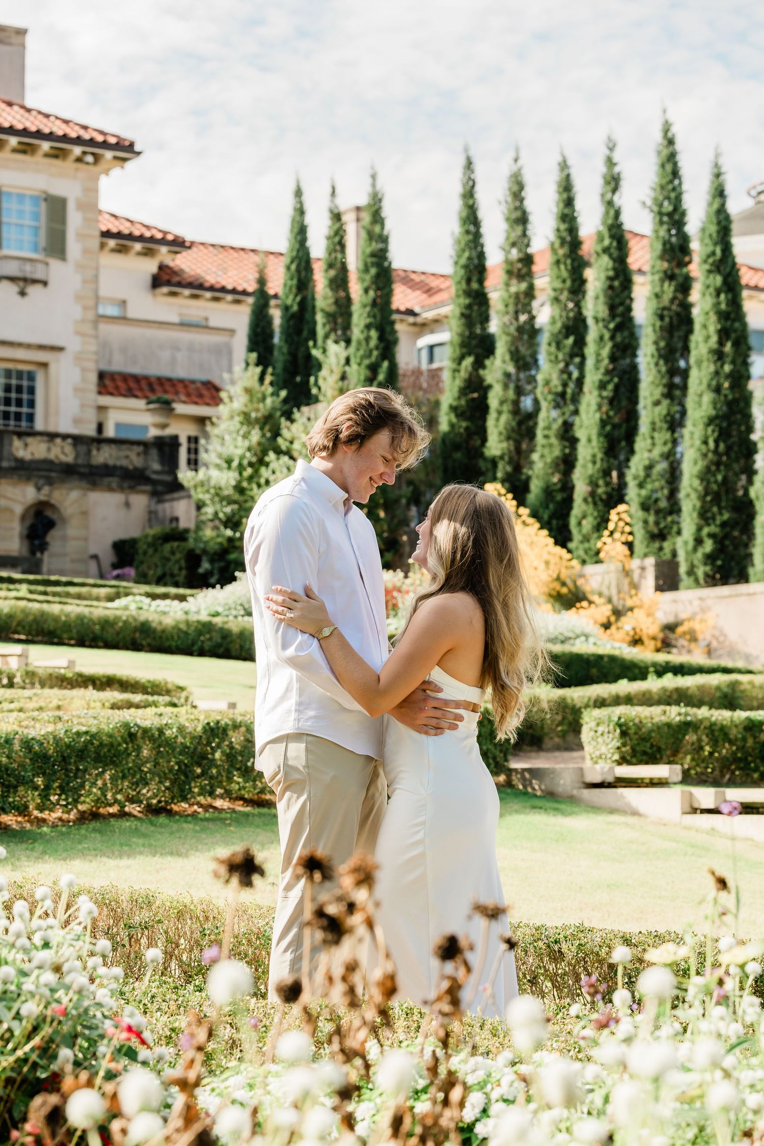 A couple shares romantic moments in a garden with cypress trees and a Mediterranean-style building in the background.