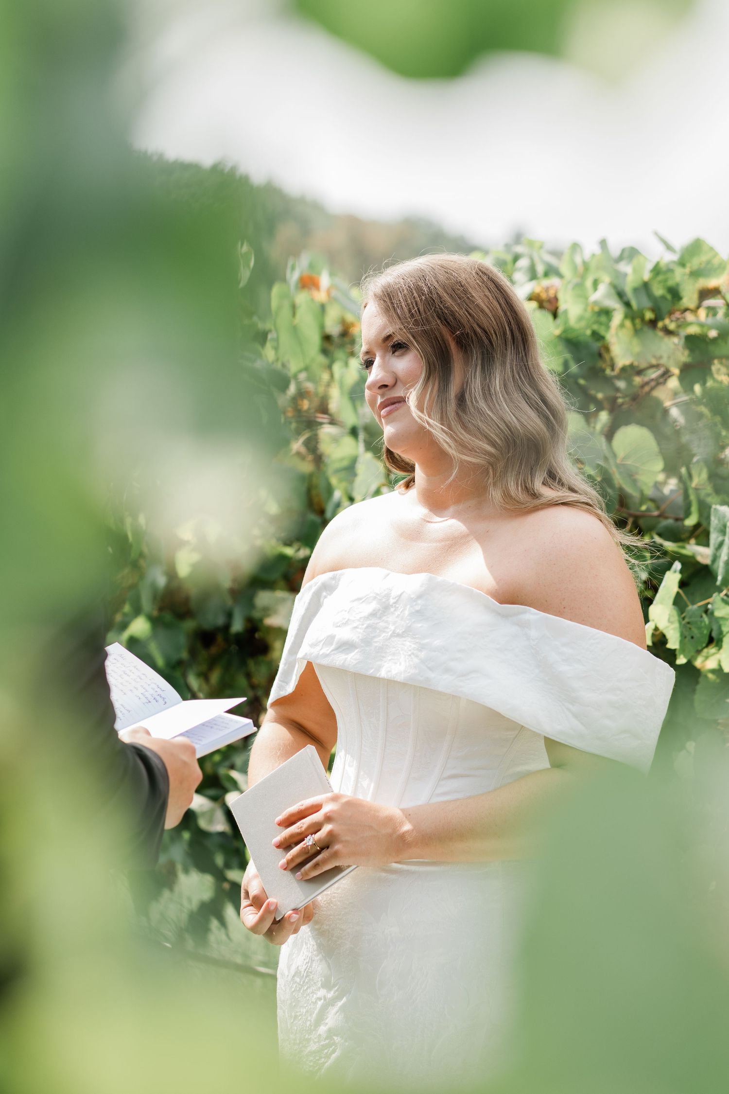 A person in a white off-shoulder dress poses gracefully among lush green grapevines in a vineyard setting.