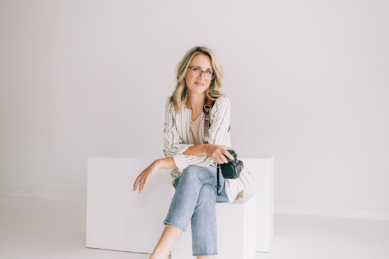A person in a white blouse and distressed jeans sits casually on a white bench in a minimalist studio setting.