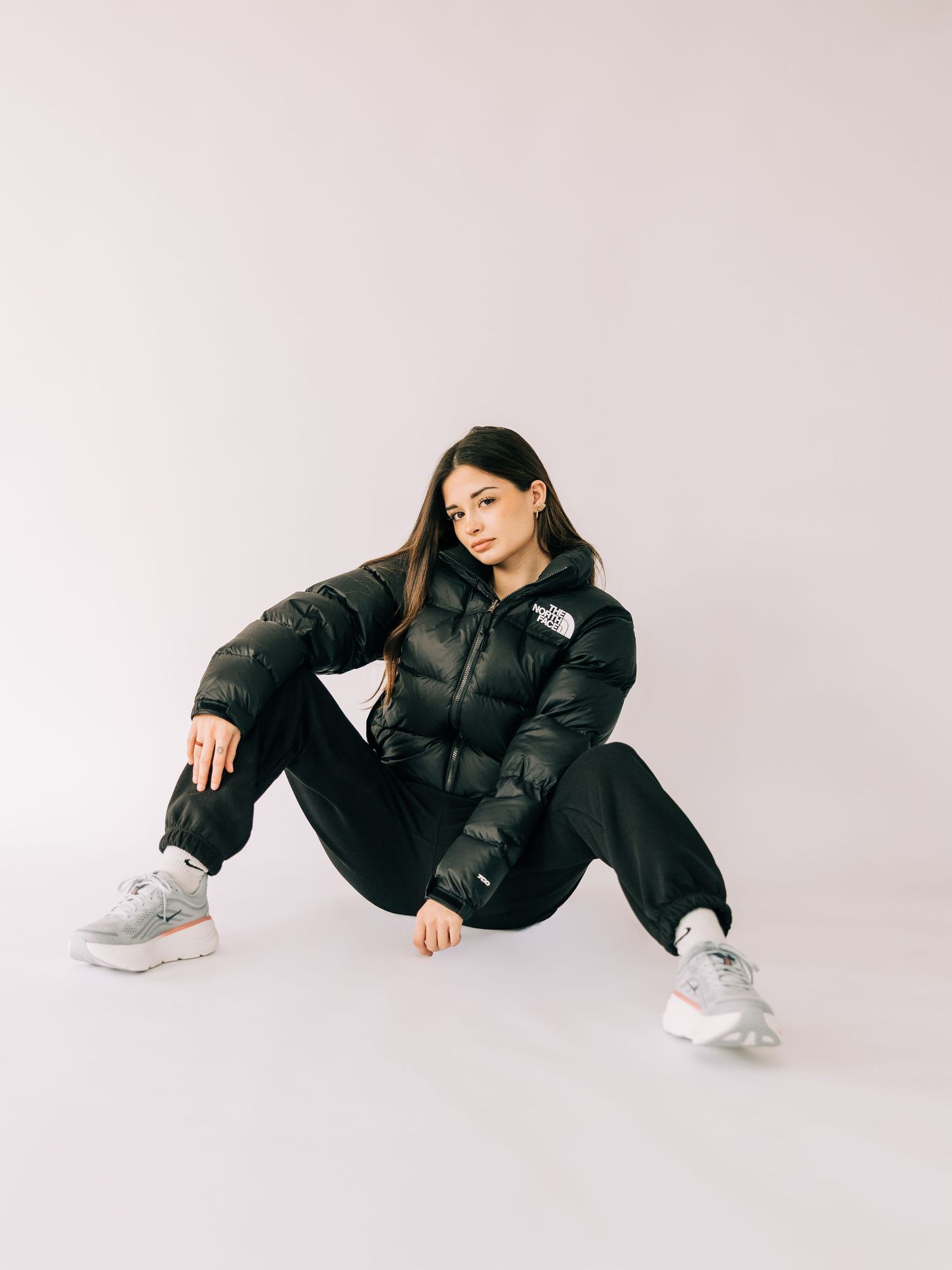 A person in a black puffer jacket and sneakers poses in a modern minimalist setting against a light gray background.