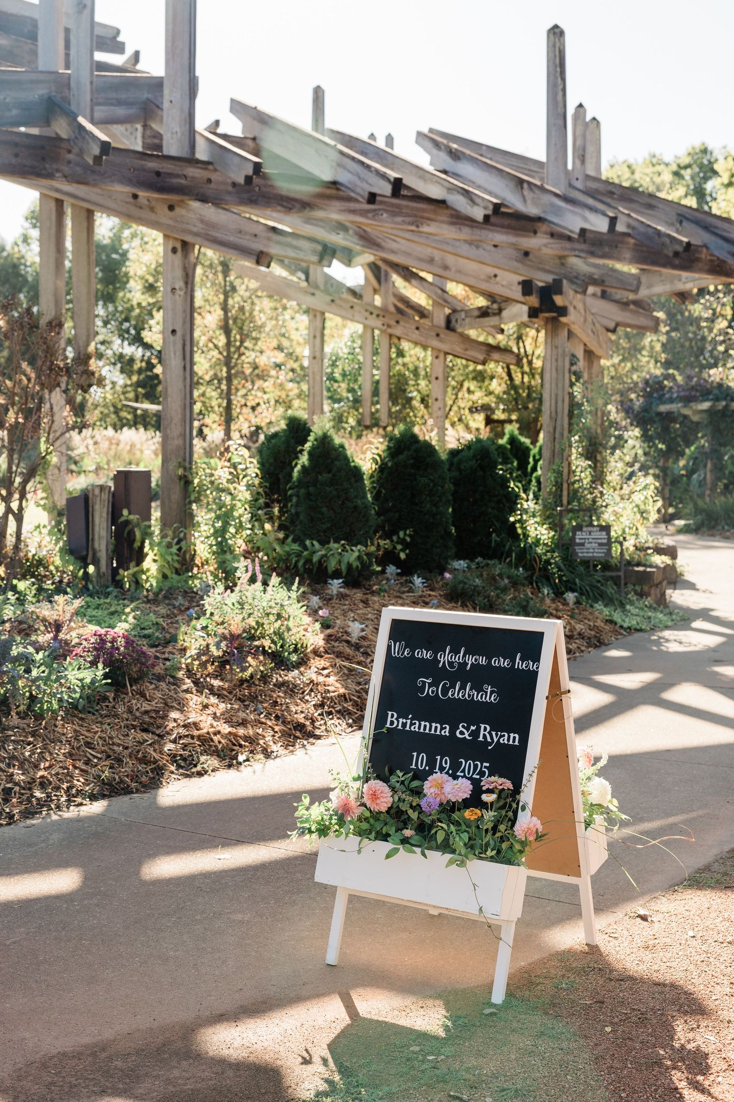 Outdoor garden walkway with wooden pergola and small chalkboard signs along a landscaped path.