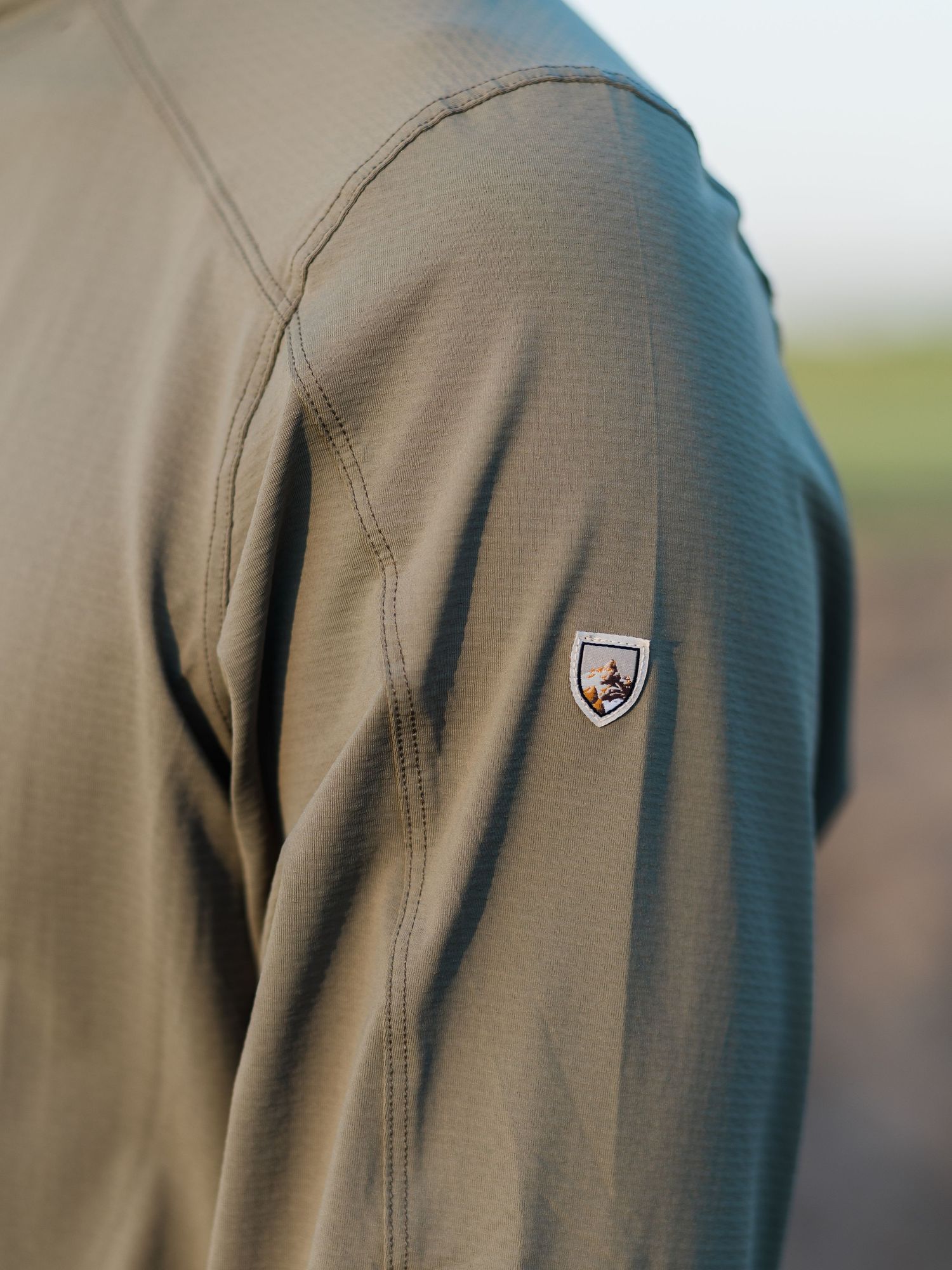 Close-up of an olive green performance jacket sleeve featuring a small shield logo patch on the shoulder.