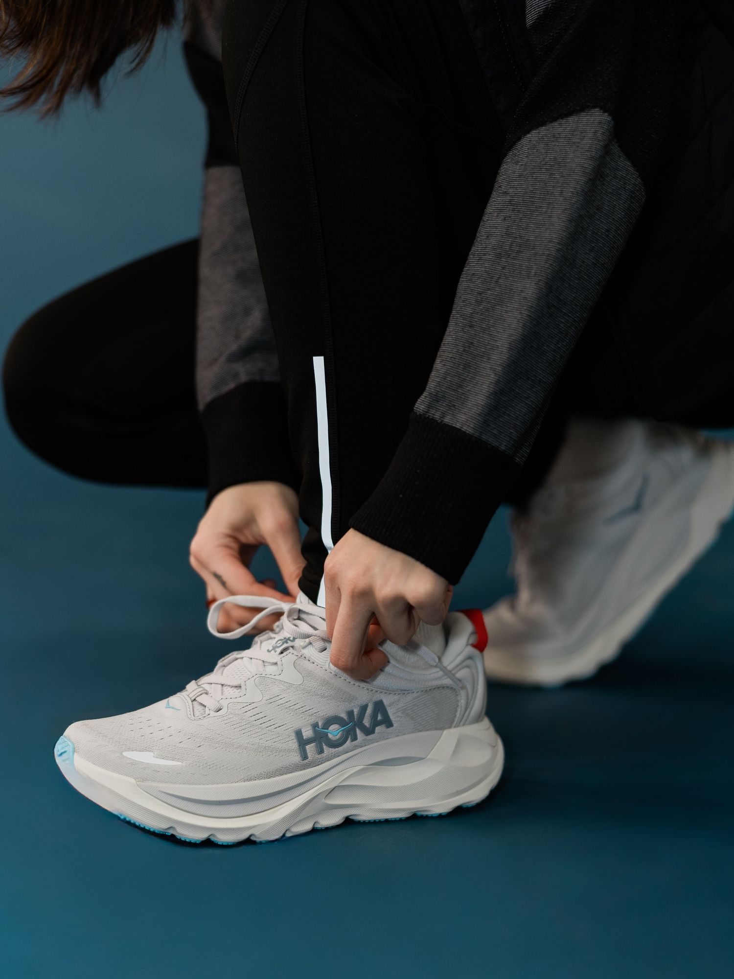 Person tying laces on white HOKA running shoes against a turquoise background.