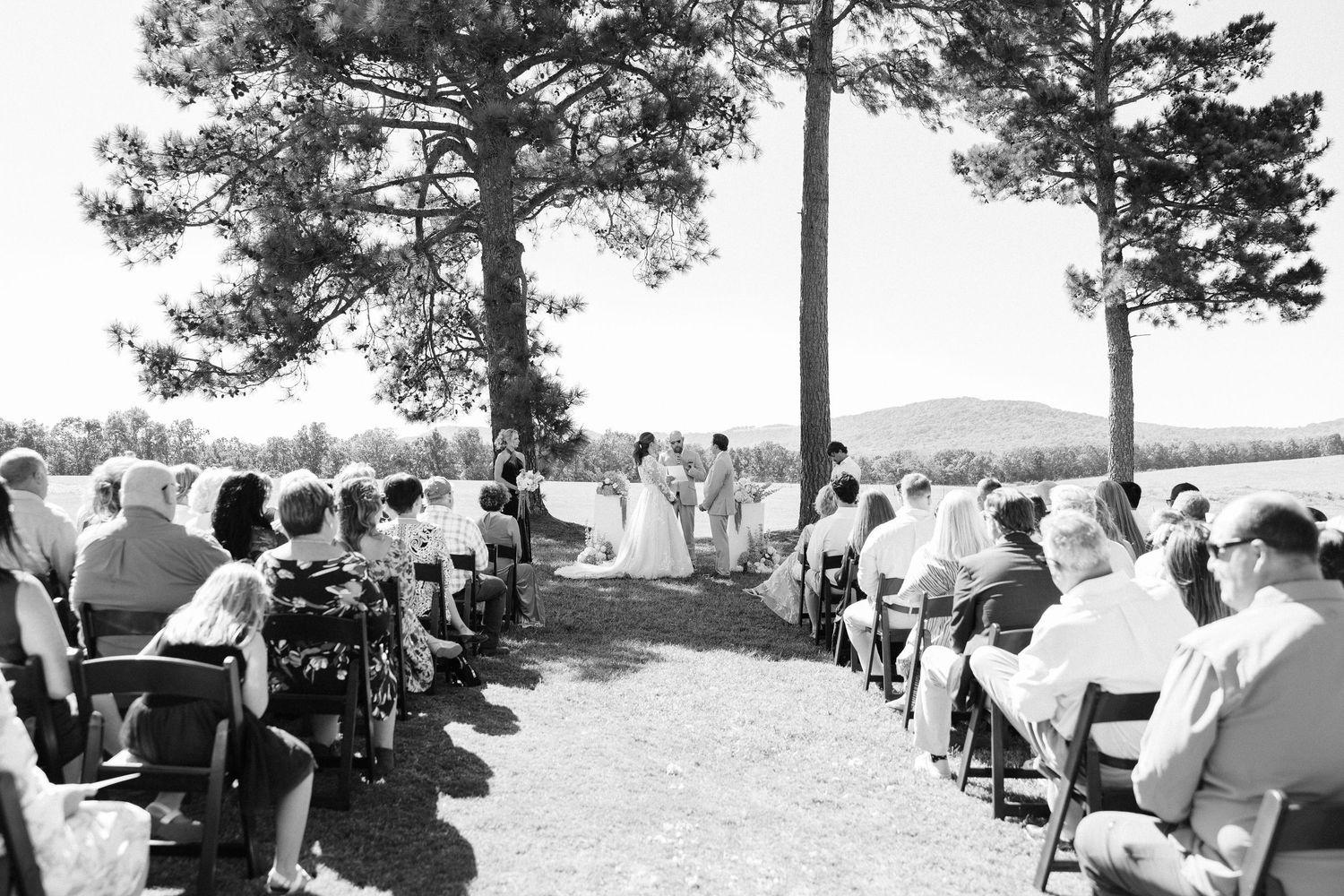 A black and white panoramic photograph shows a large group of people gathered outdoors among tall pine trees.