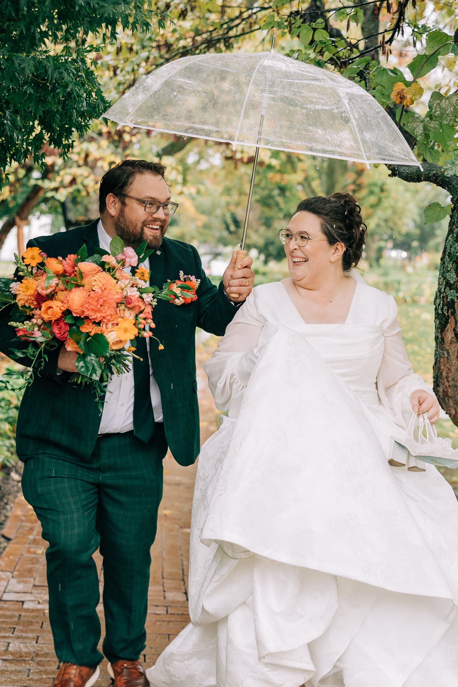 A rainy day wedding photoshoot with clear umbrellas and orange floral bouquets in a garden setting.