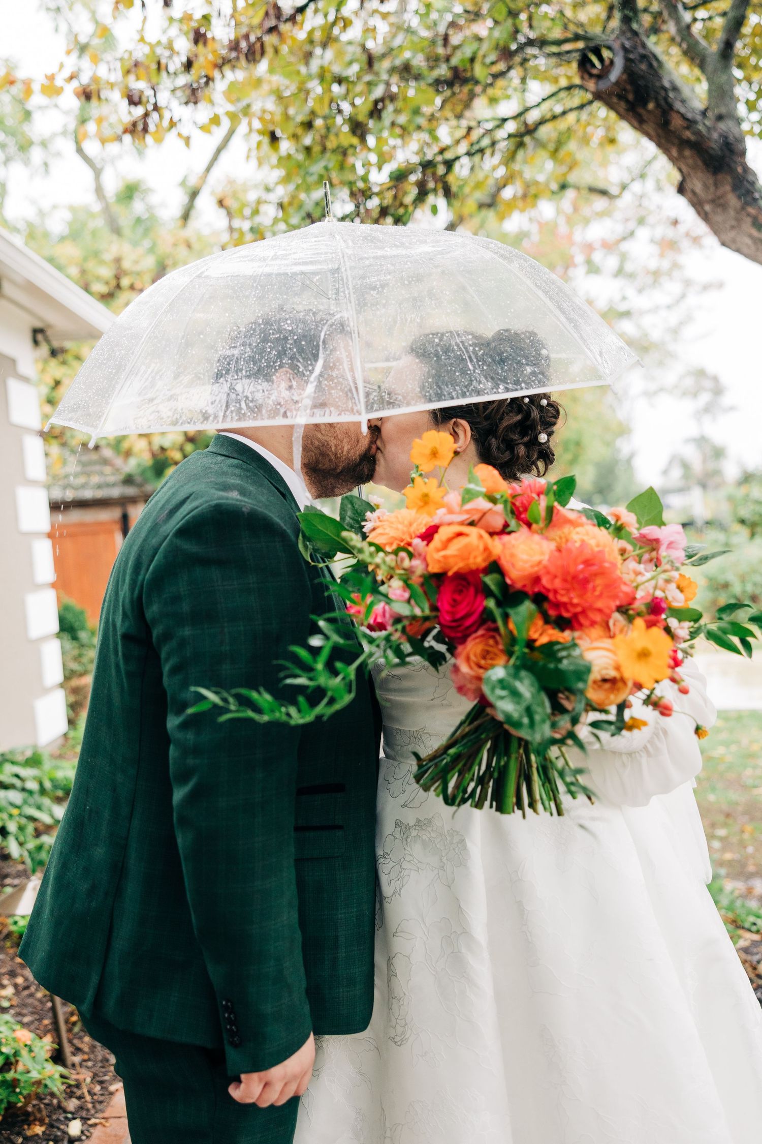 A couple shares an intimate moment under a clear umbrella with vibrant coral and orange wedding bouquet.