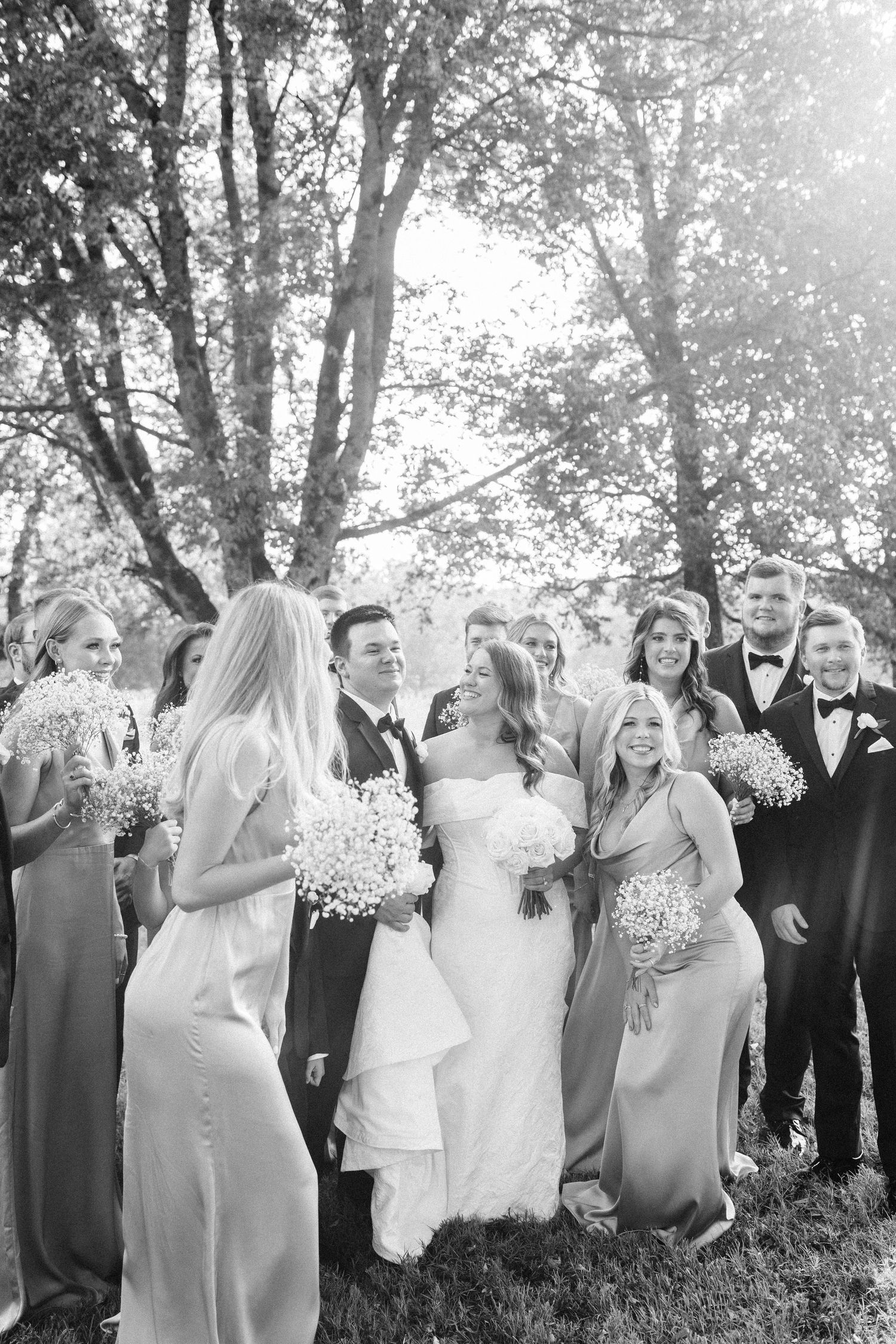 A wedding party in formal attire stands together outdoors with bridesmaids in light dresses and groomsmen in dark suits.