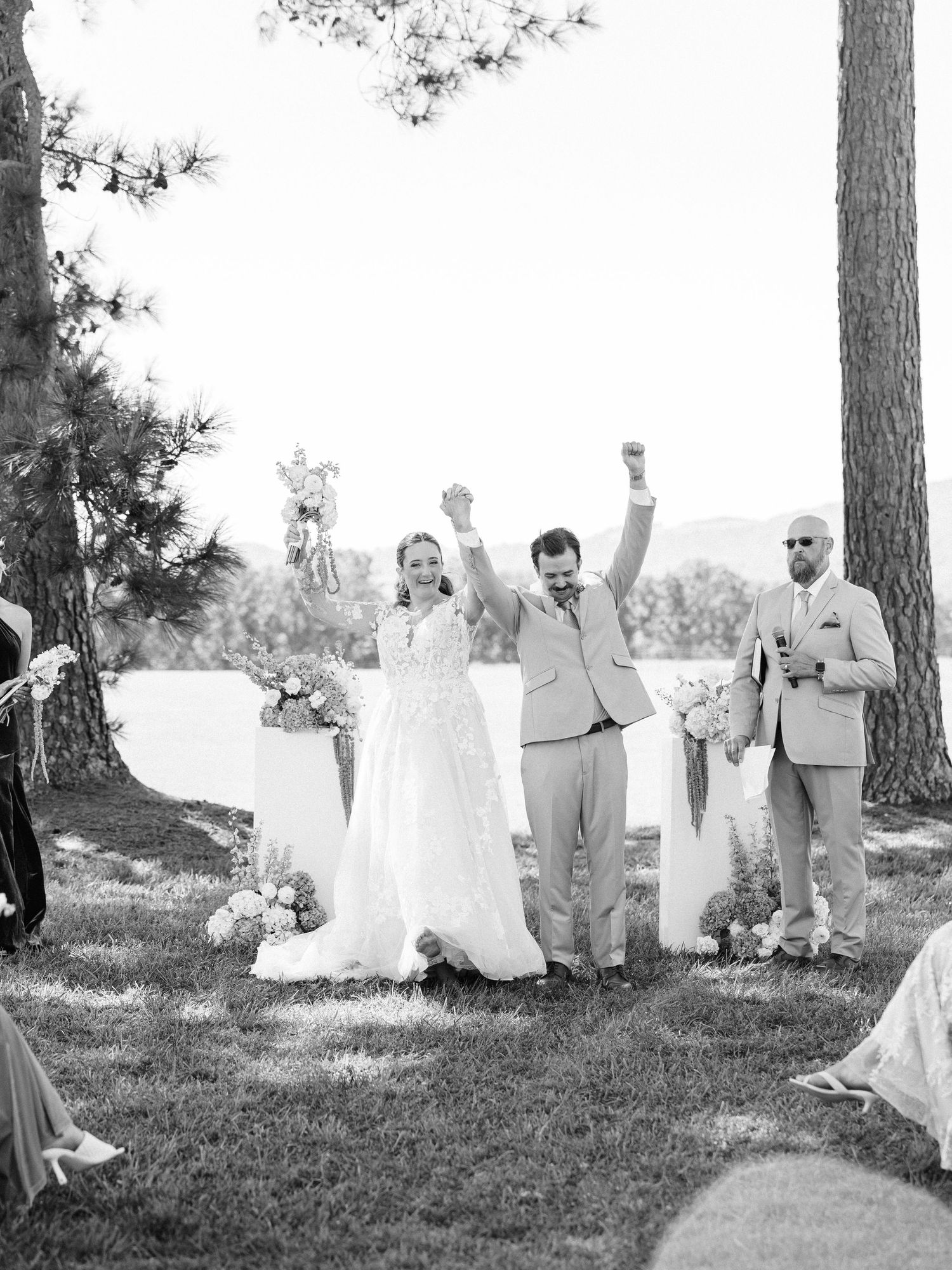 Joyful wedding party celebrating with raised arms under tall pine trees.