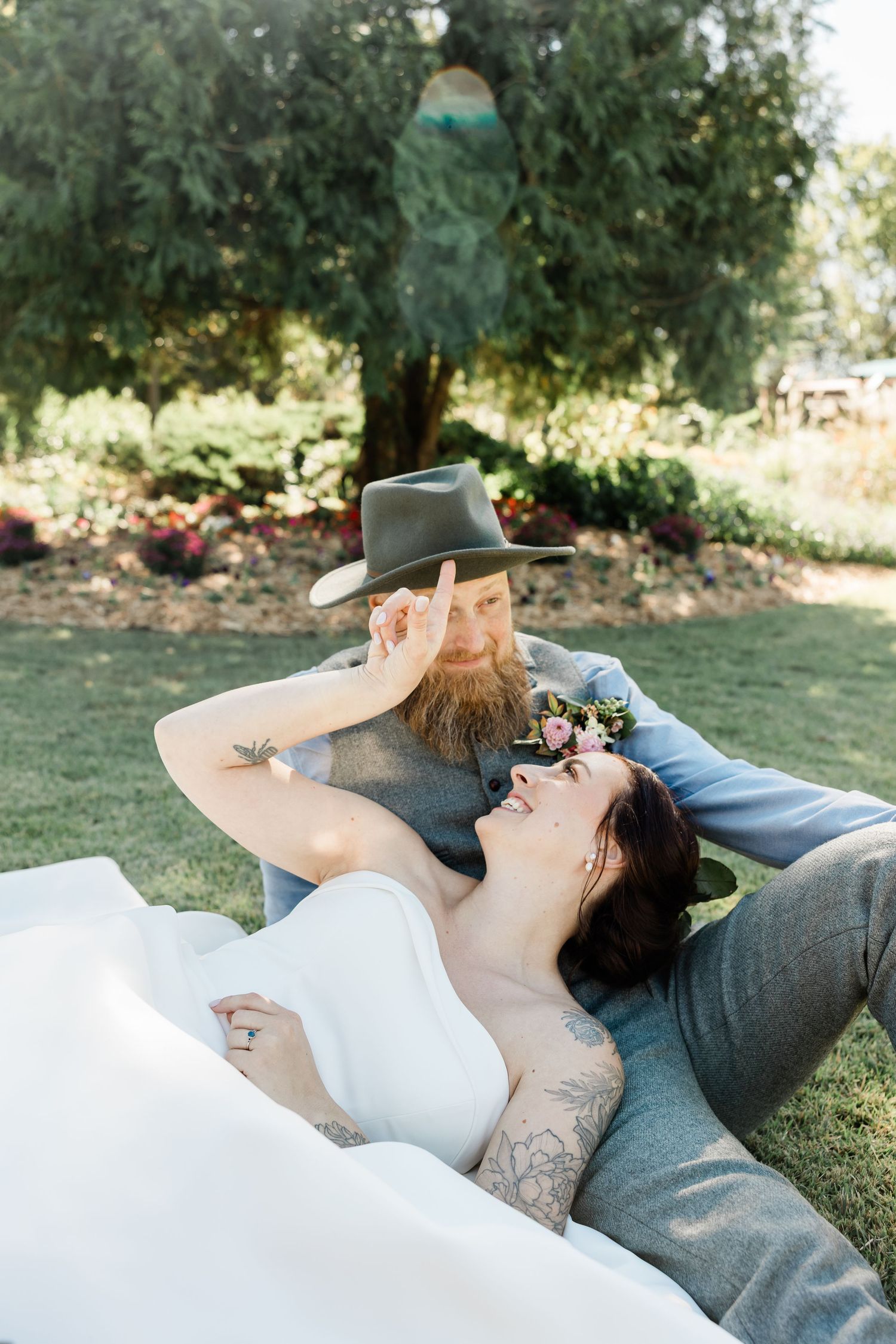 A couple lying on grass in an outdoor garden setting with flowers and greenery in the background.