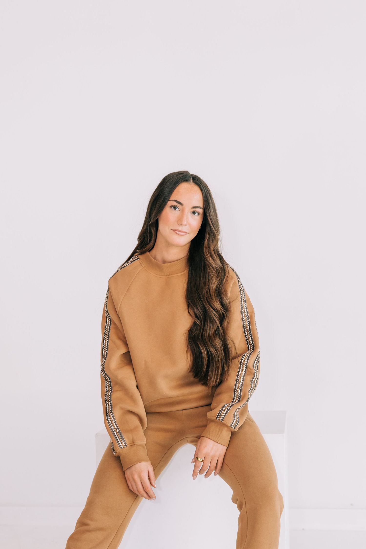 Woman in camel brown matching sweatsuit with stripe detail, sitting casually against white background.