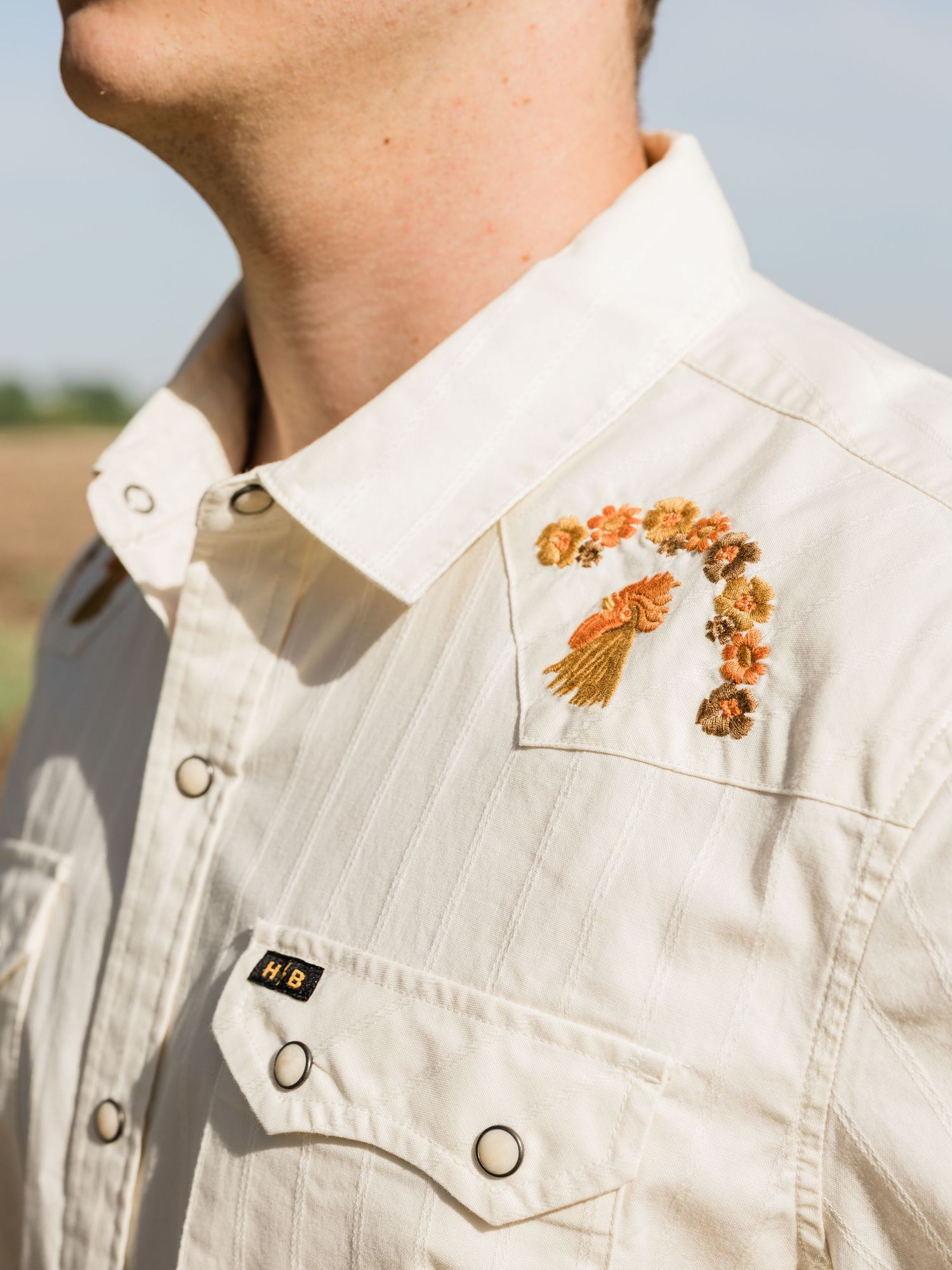 Close-up of a cream western shirt with floral embroidery and snap buttons, worn by a man outdoors.