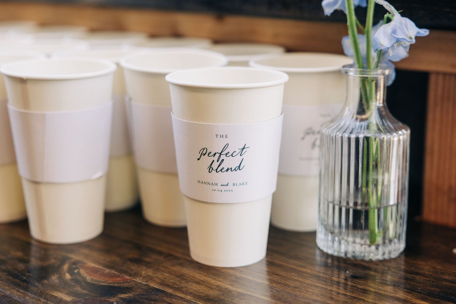 Row of white paper cups and glass vases with flowers on a wooden table surface.