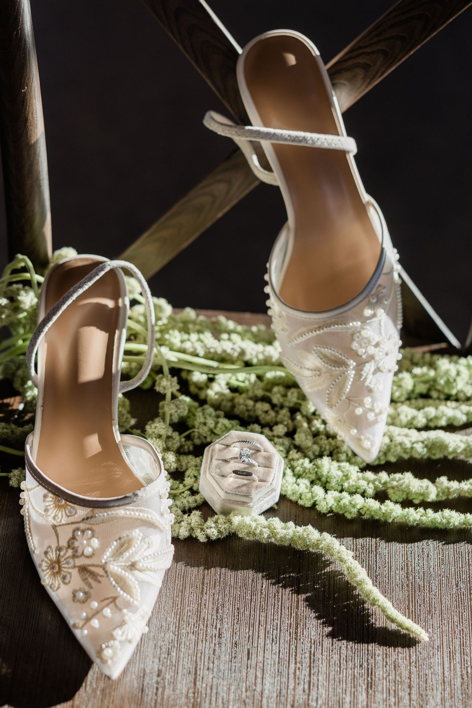 Delicate white bridal shoes with floral lace details and pearls displayed against a rustic wooden surface.
