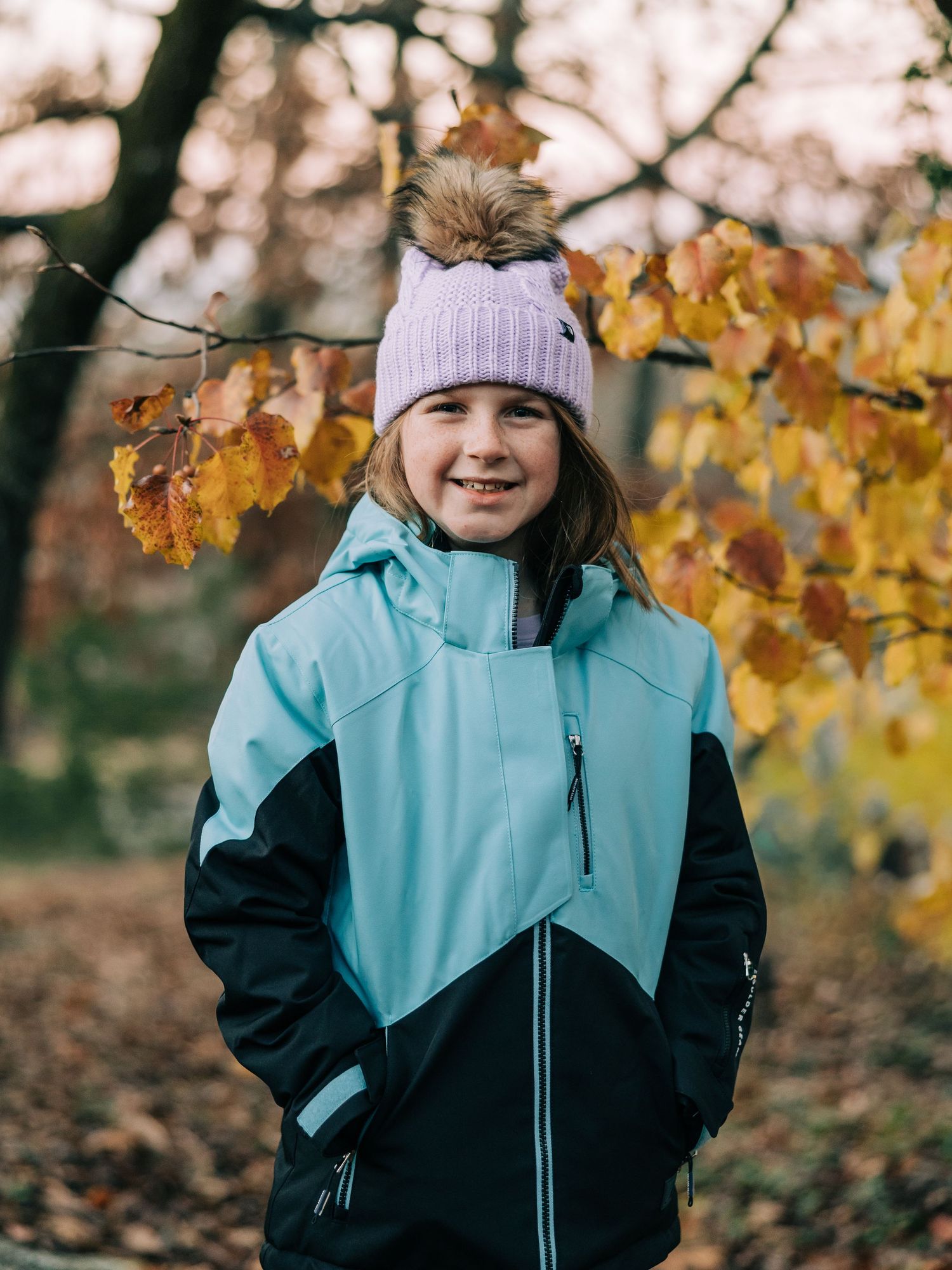 A child wearing a purple knit hat and turquoise winter coat stands among golden autumn leaves in a forest setting.