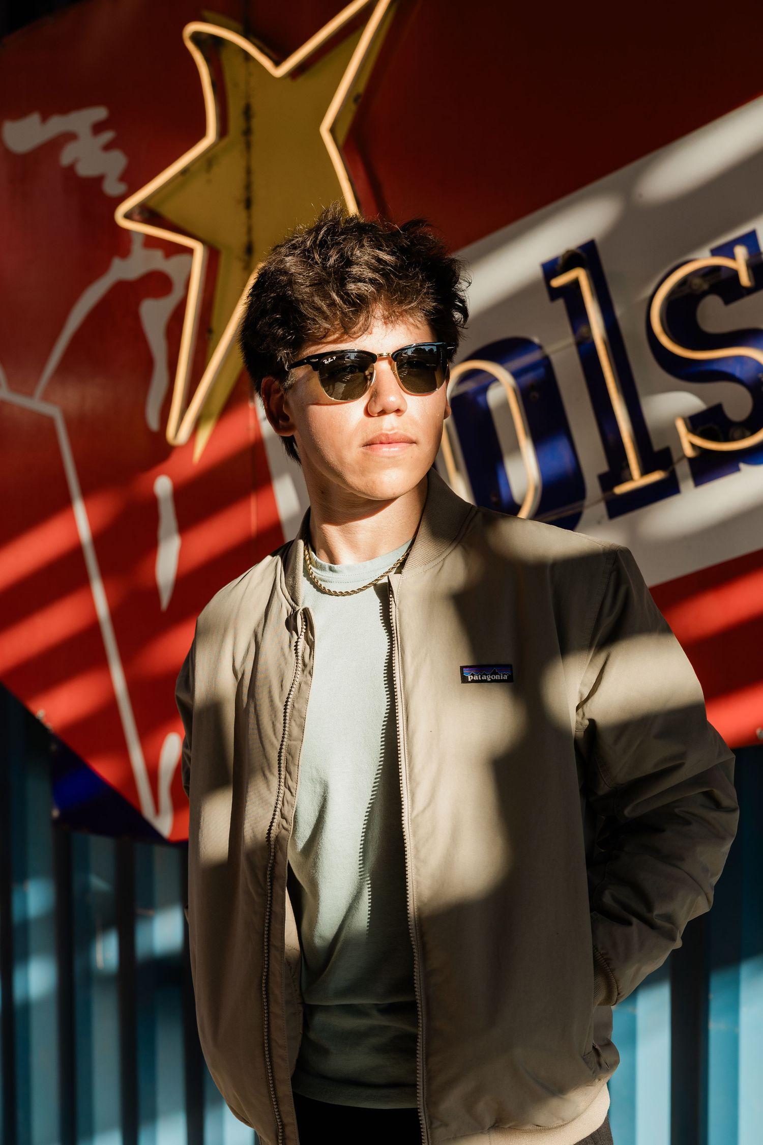 A person wearing sunglasses and a beige jacket poses against a dark background with red and blue lighting effects.