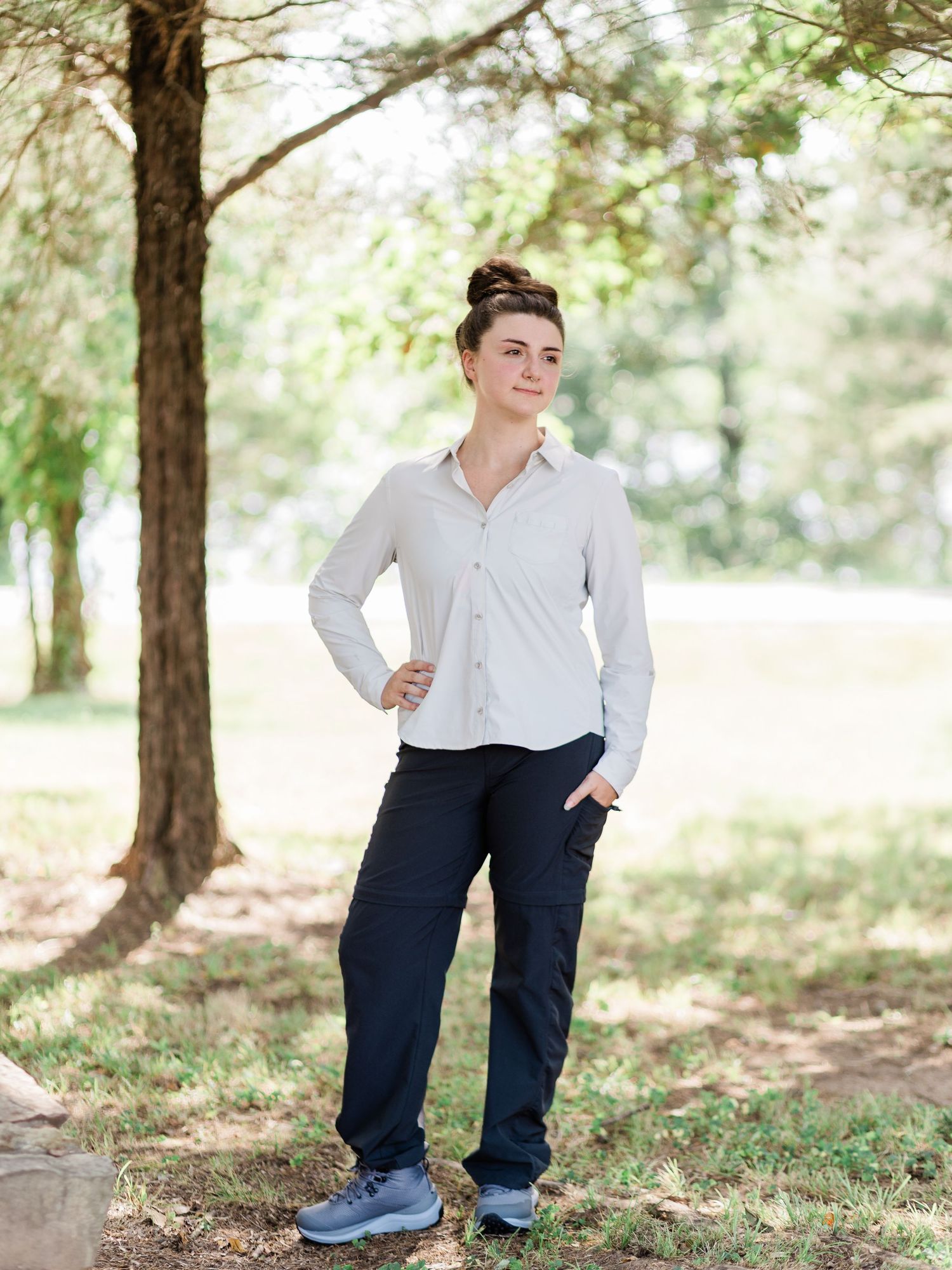 Woman in beige button-up shirt and navy cargo pants standing outdoors under trees in a park setting.