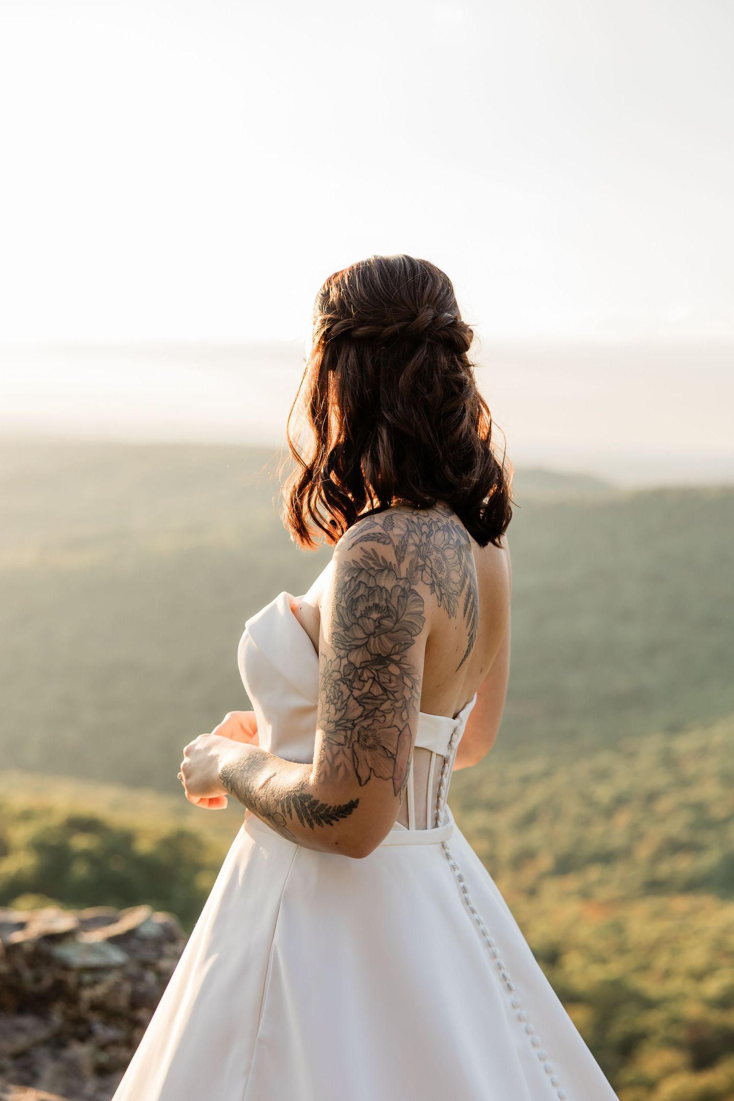 Bride in white dress with tattoos stands looking out over mountain vista.