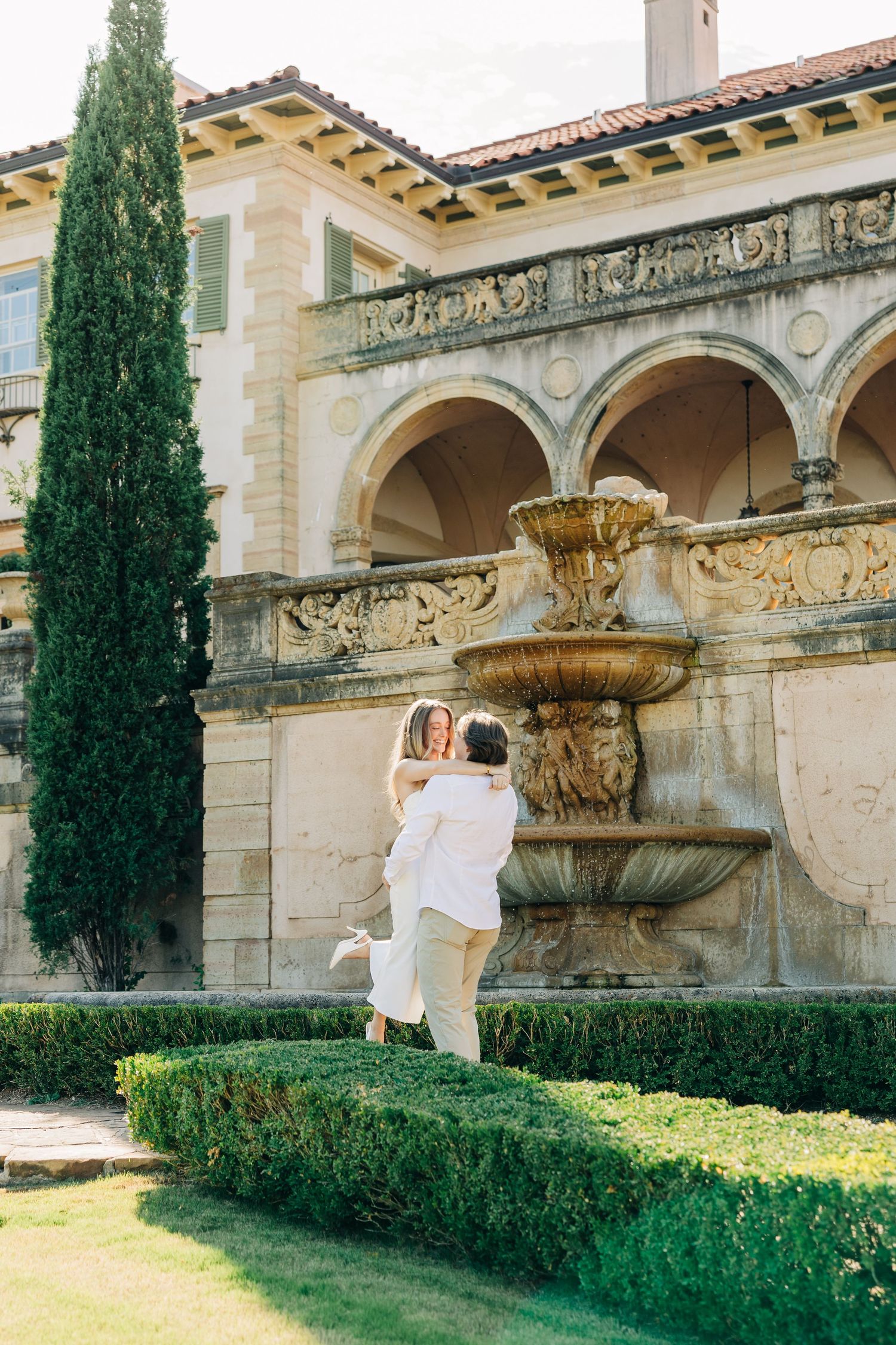 Elegant stone archways and cypress trees line the exterior of a historic Mediterranean-style building with manicured gardens.