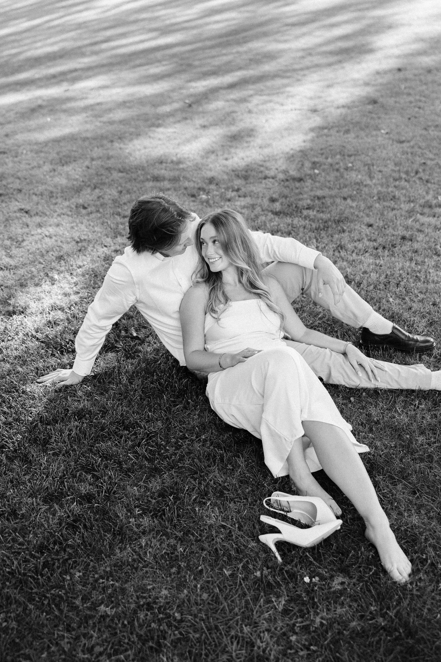 A romantic black and white photo of a couple lounging together on grass during a sunny day.