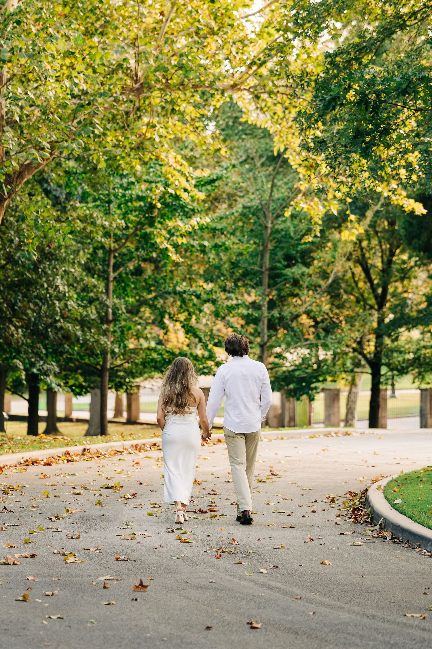 A couple walks together on a tree-lined path covered in fallen autumn leaves on a sunny day.