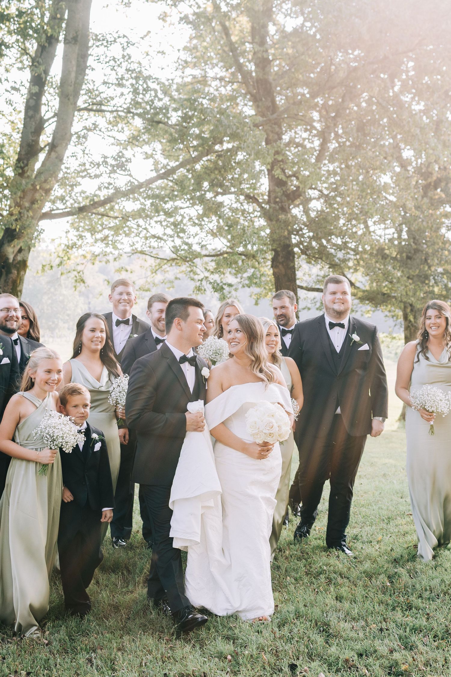 Wedding party laughs together in an outdoor setting with trees and natural sunlight creating a joyful atmosphere.