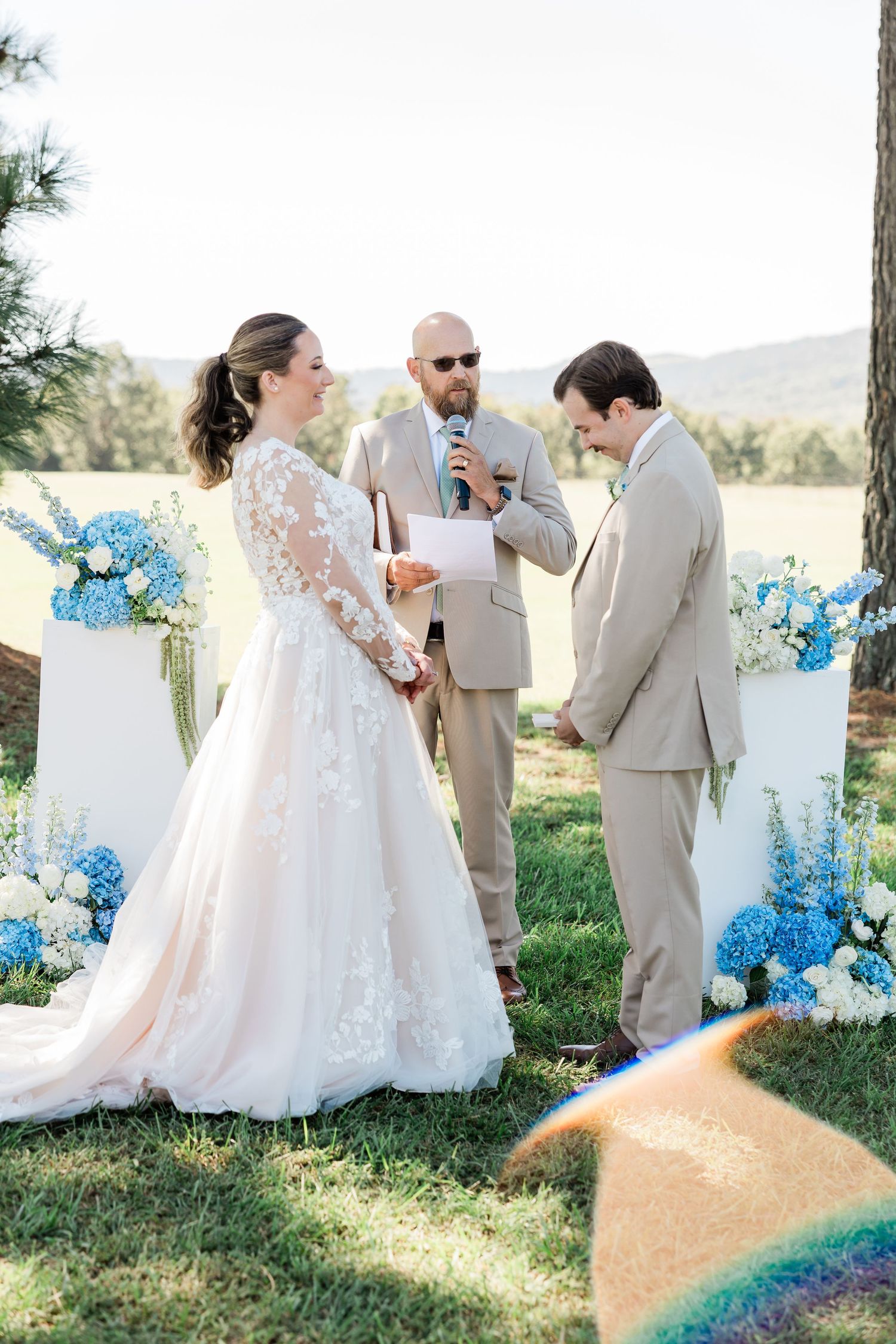 Wedding party in beige suits and white dress gathered under pine trees with blue floral accents.