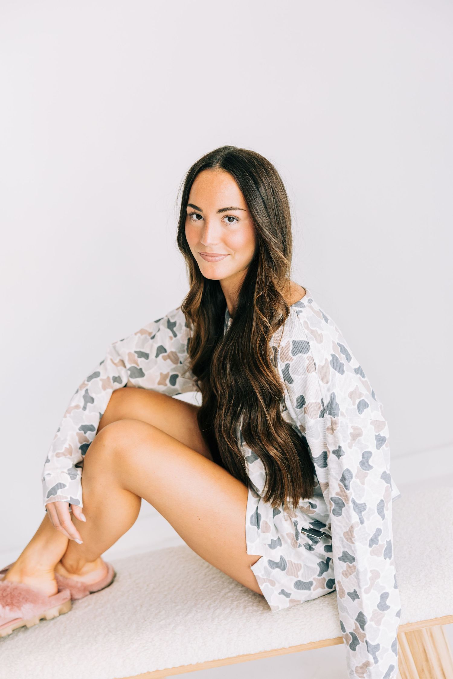 Young woman with long dark hair sitting on a bed wearing a floral robe and pink slippers, smiling at camera.