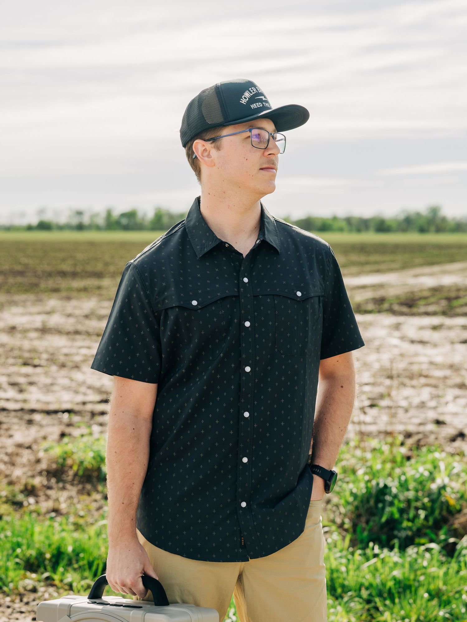 Young man in black patterned shirt and cap standing in a farm field on a cloudy day.