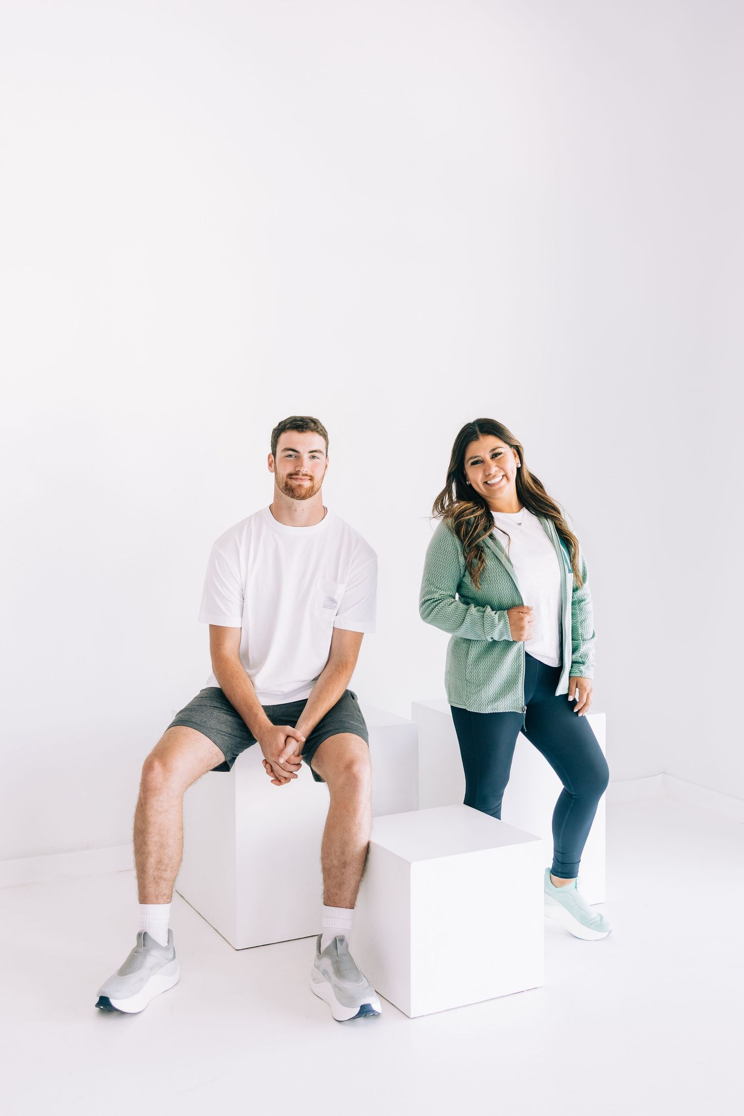 A man and woman in casual athletic wear pose on white cubes against a white background, smiling at the camera.