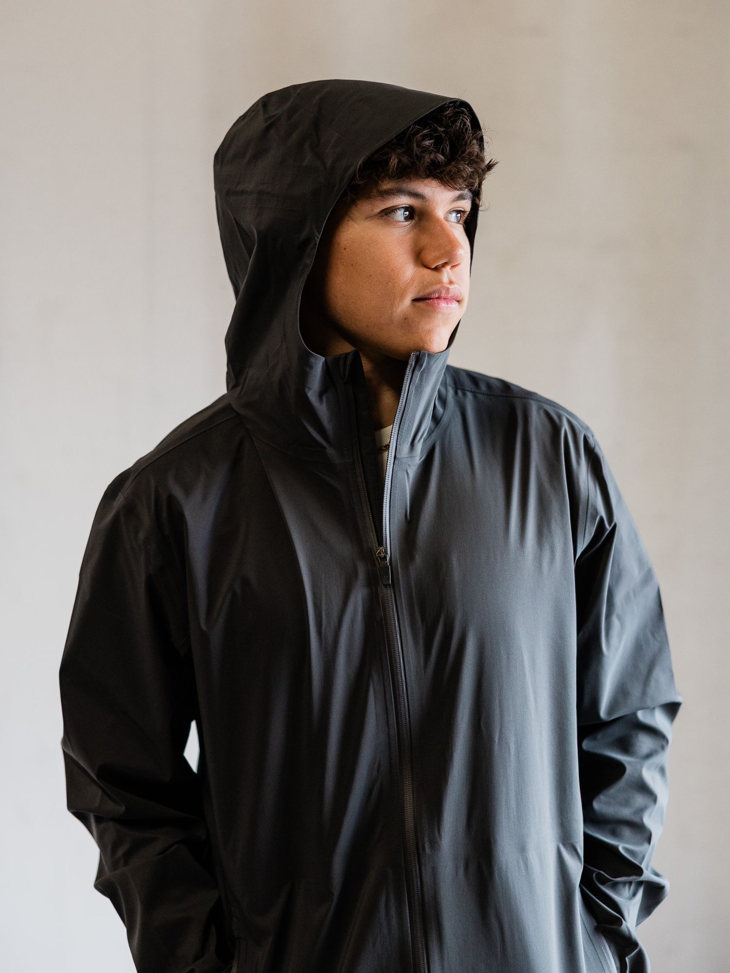Young man wearing a black hooded waterproof zip-up jacket, posing against a light gray background.