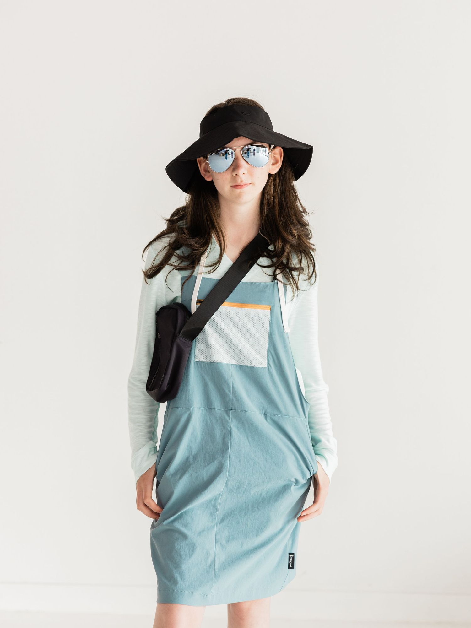 Woman wearing a light blue colorblock dress over a white long-sleeve shirt, black bucket hat, sunglasses, and crossbody bag.