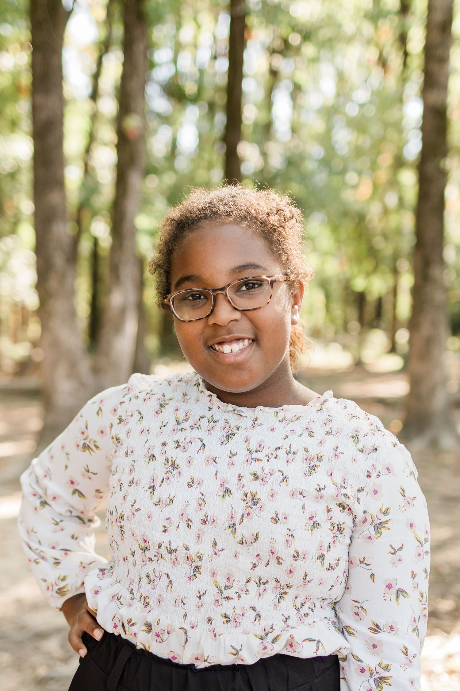 A person in glasses and a white floral top poses outdoors in a wooded setting with natural lighting.