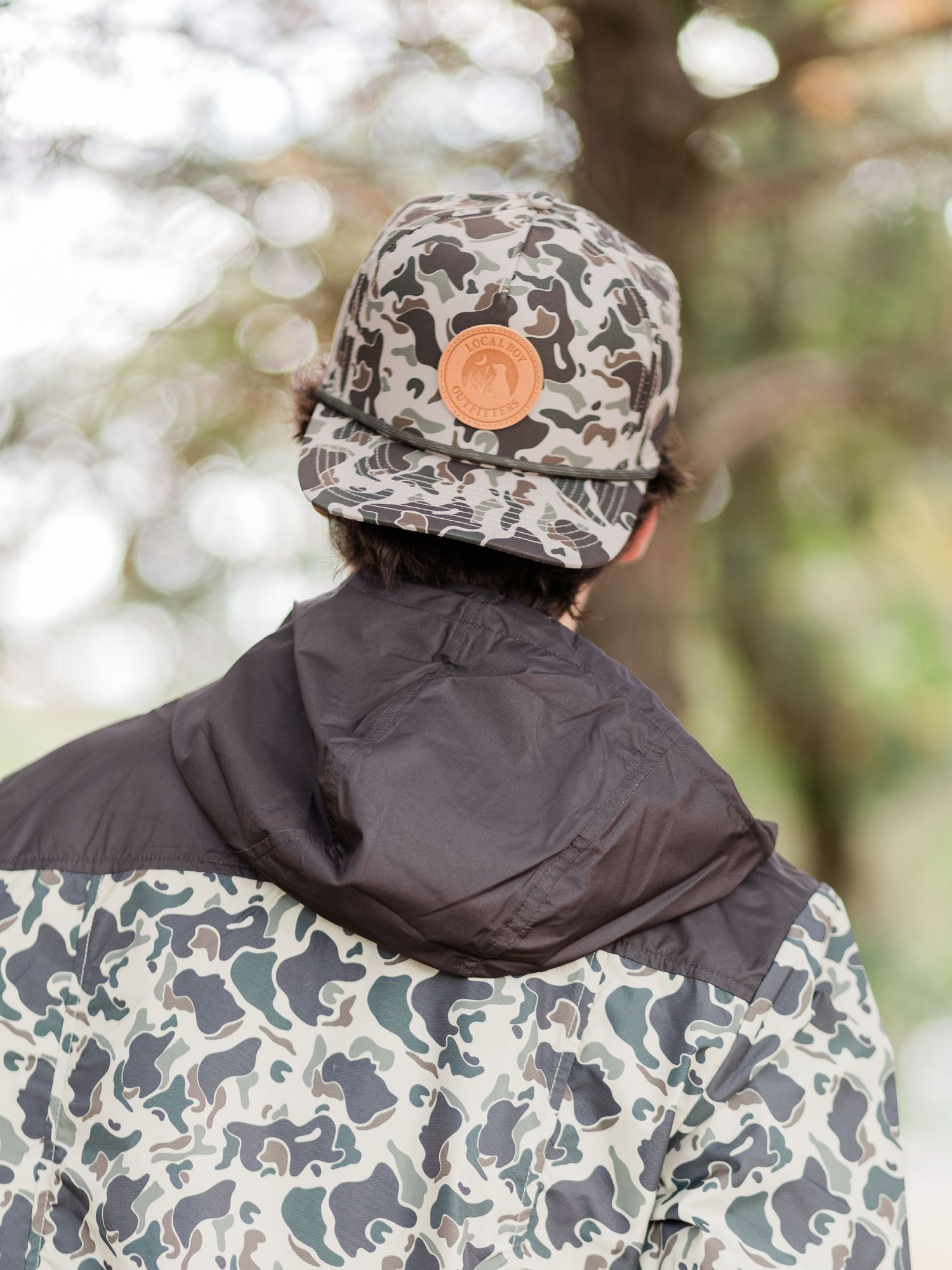 Person wearing camo bucket hat with leather patch and matching camo jacket, viewed from behind outdoors.