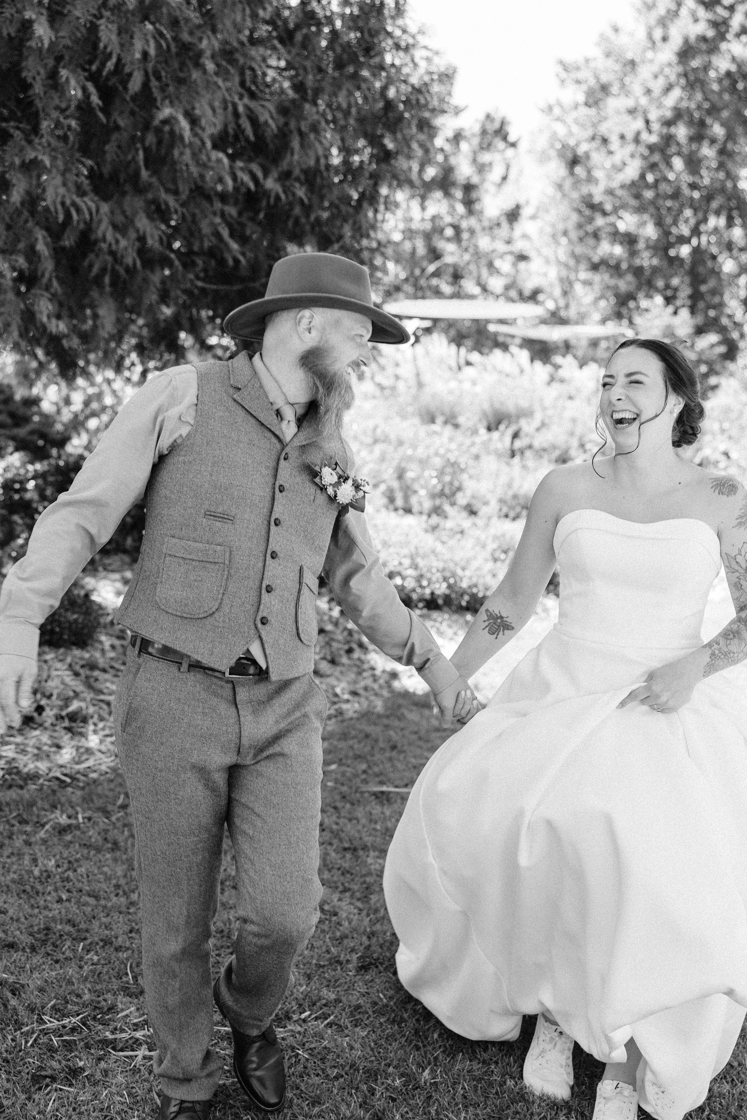 A couple takes playful wedding photos outdoors with the bride in a white dress and groom in casual attire.