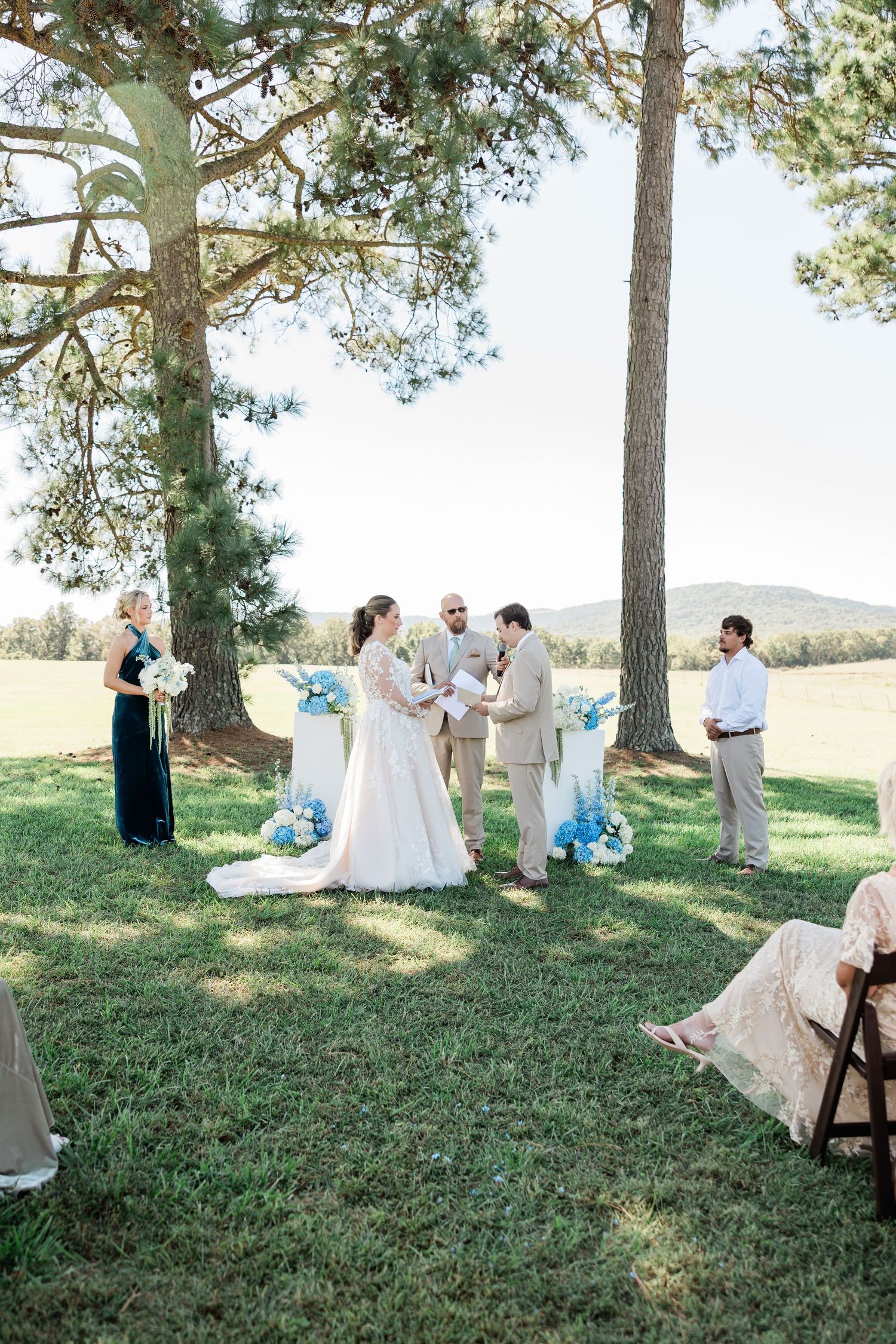 Wedding ceremony setup under pine trees with white chairs and blue floral decorations.