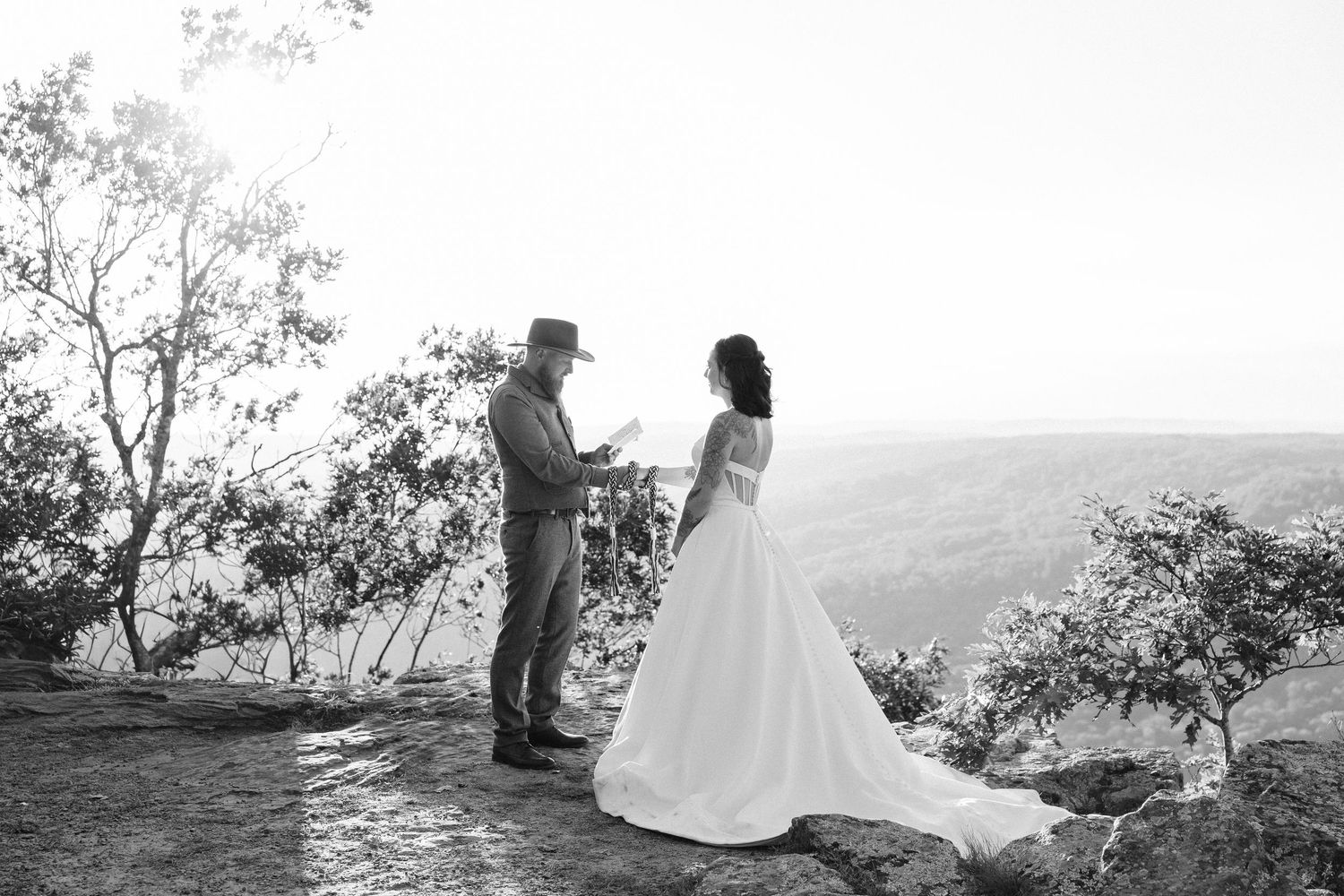 A wedding couple stands on a rocky overlook with mountains in the distance during sunset.