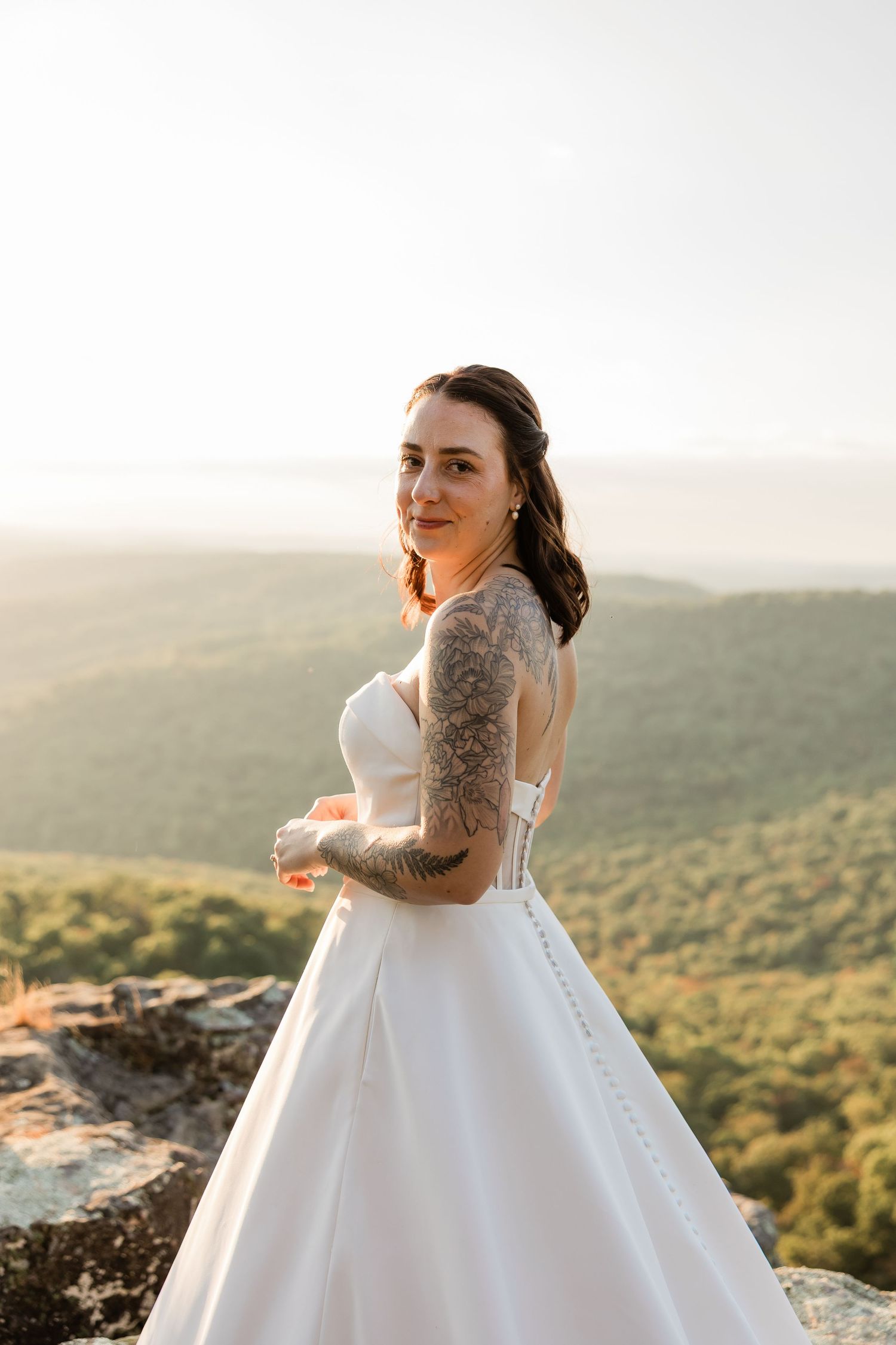 A bride in a flowing white ballgown poses against a scenic mountain backdrop at golden hour.