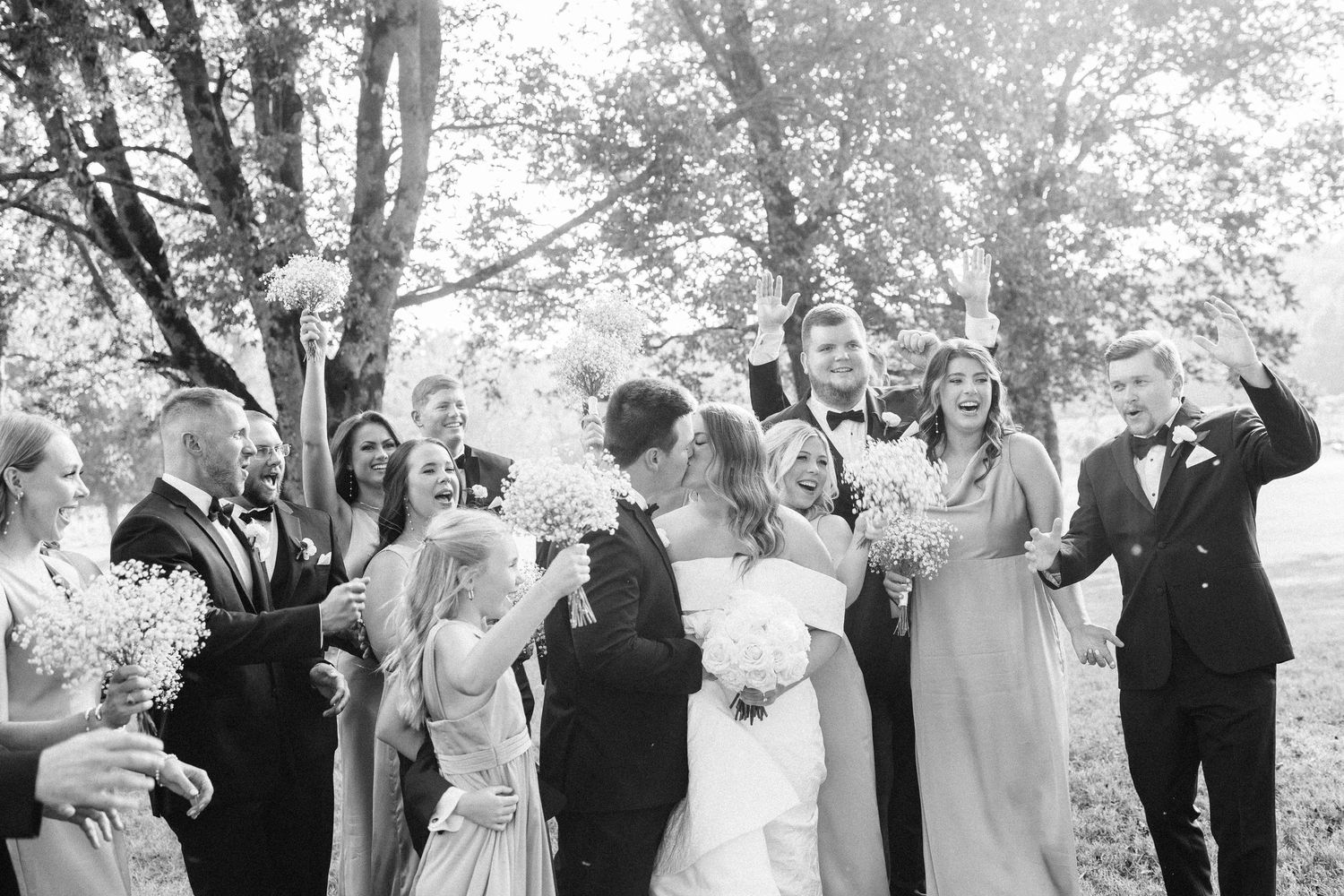 A wedding party laughs together while posing outdoors under trees in a candid black and white photograph.
