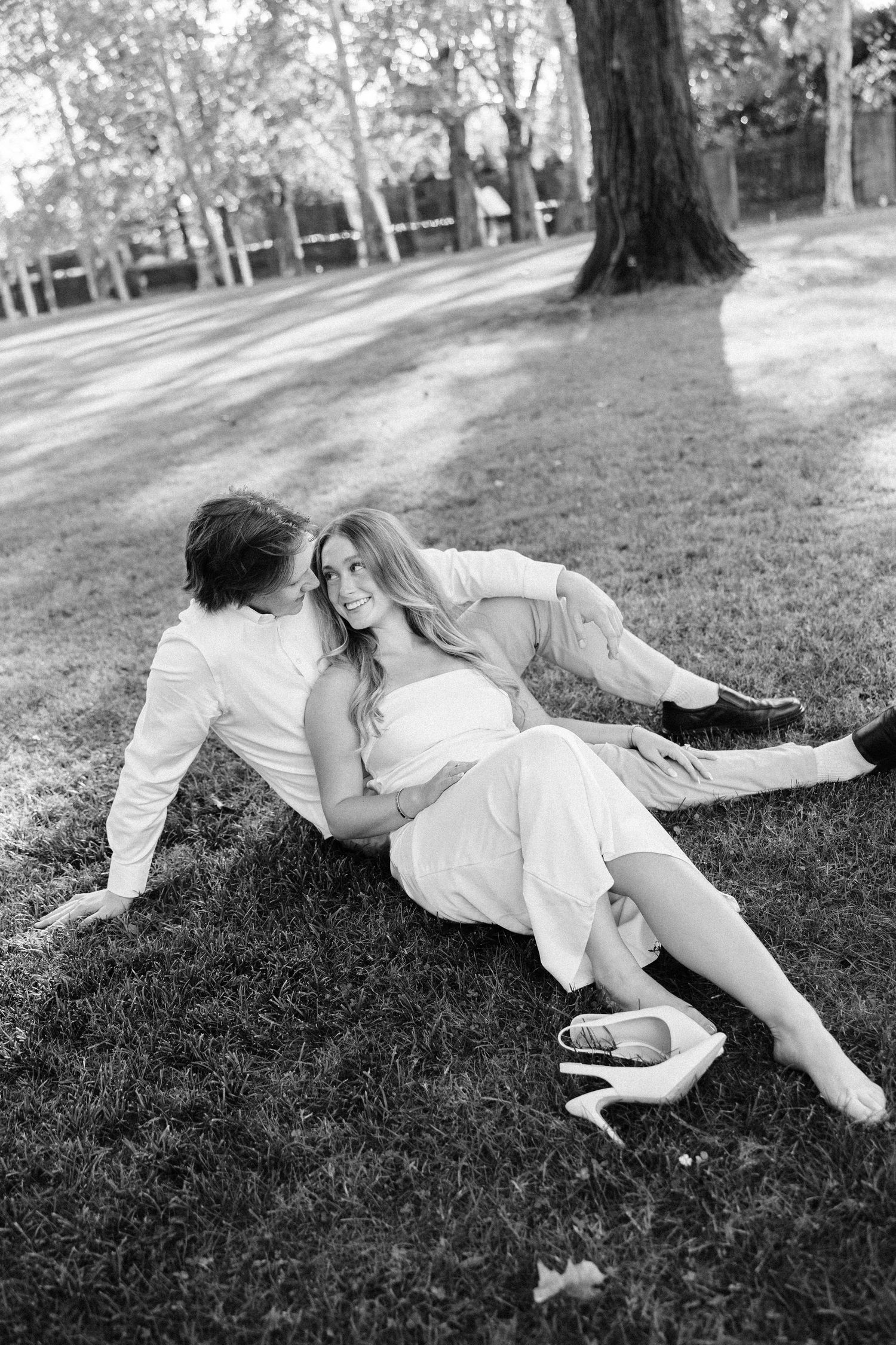 A couple in white clothing lies playfully on green grass in a park setting.