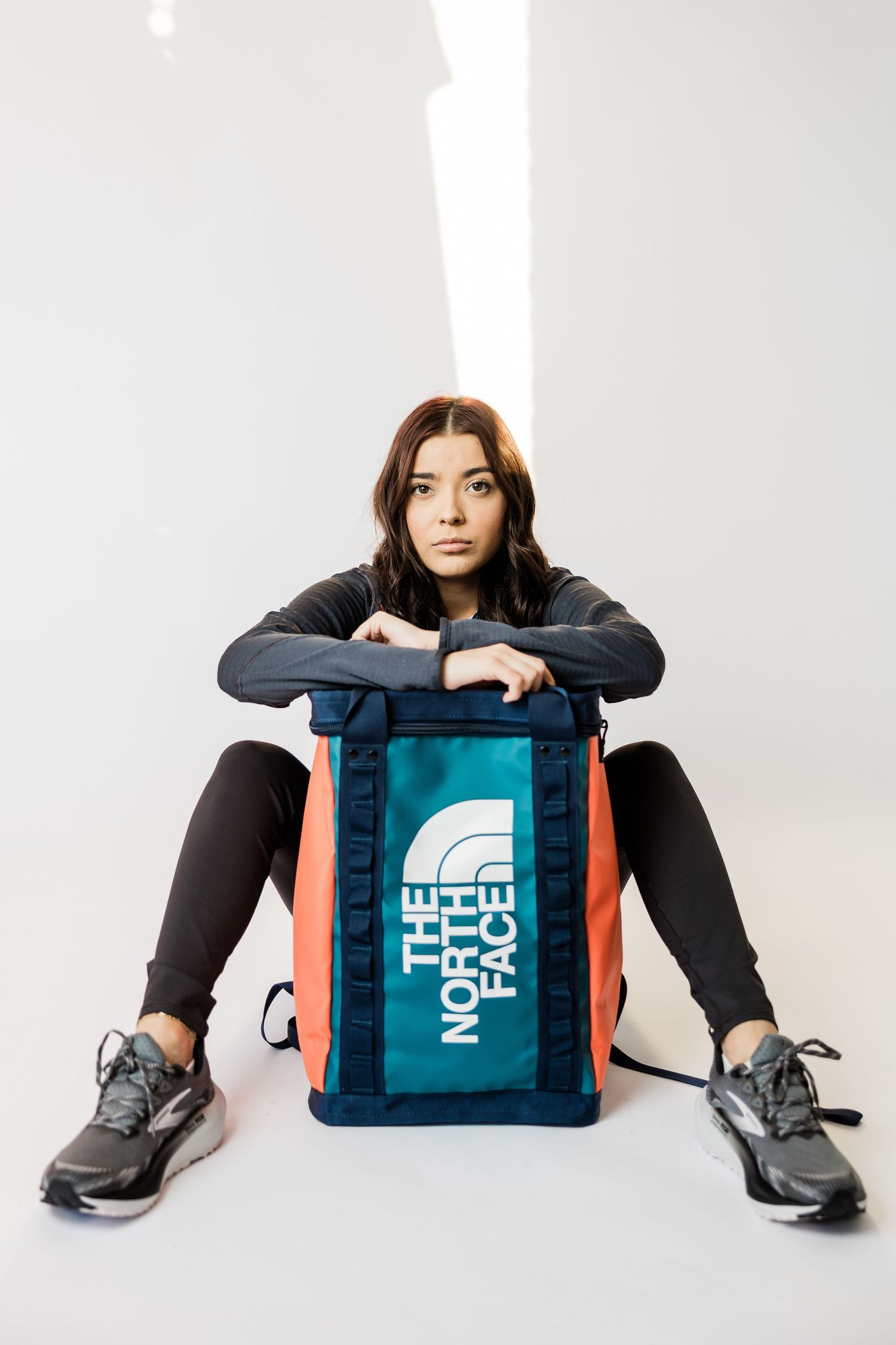 Person sits on white floor leaning on teal North Face backpack while wearing black outfit and gray sneakers.