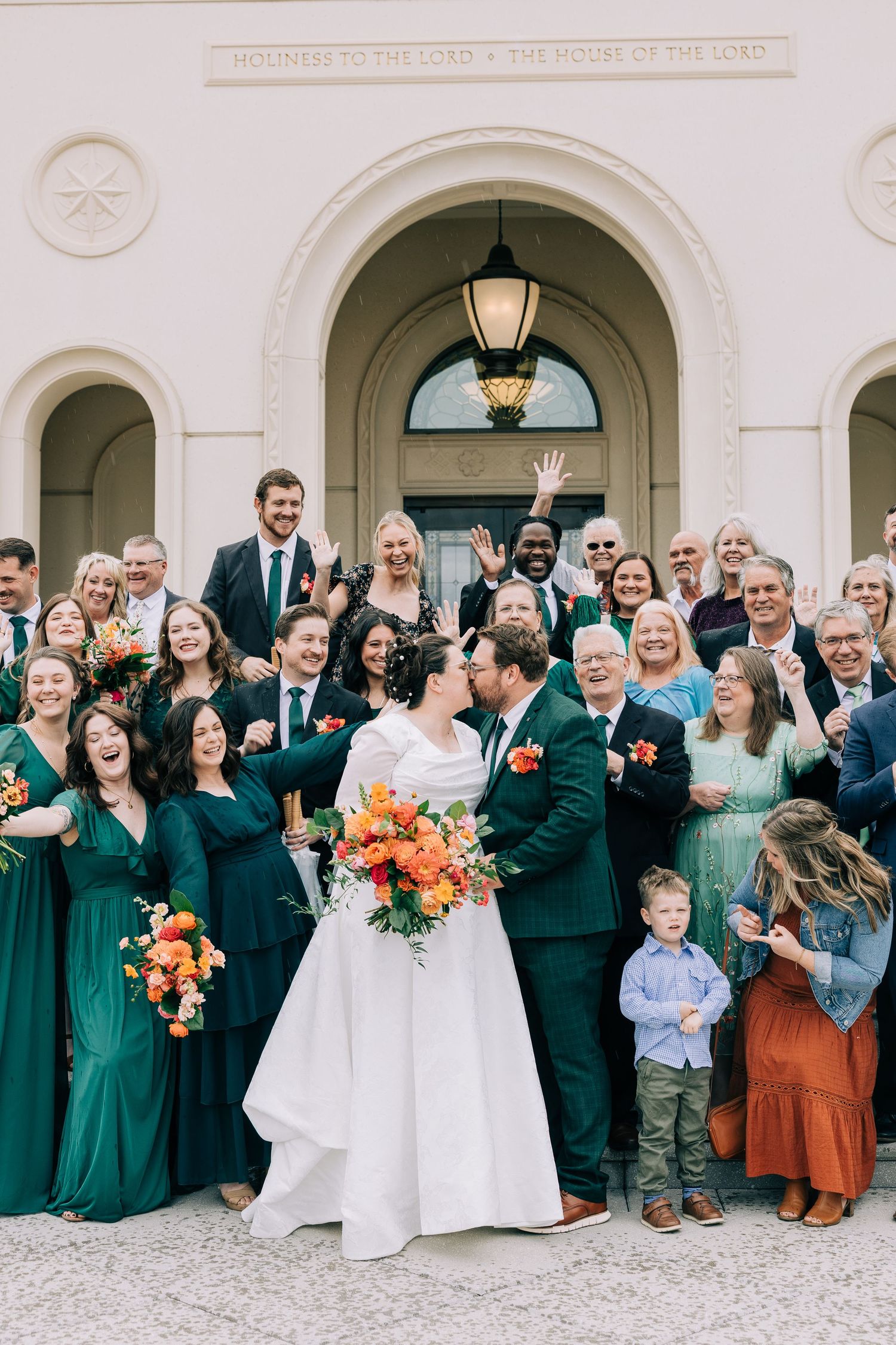 Large wedding party in emerald green dresses gathers for a group photo outside a classic building.