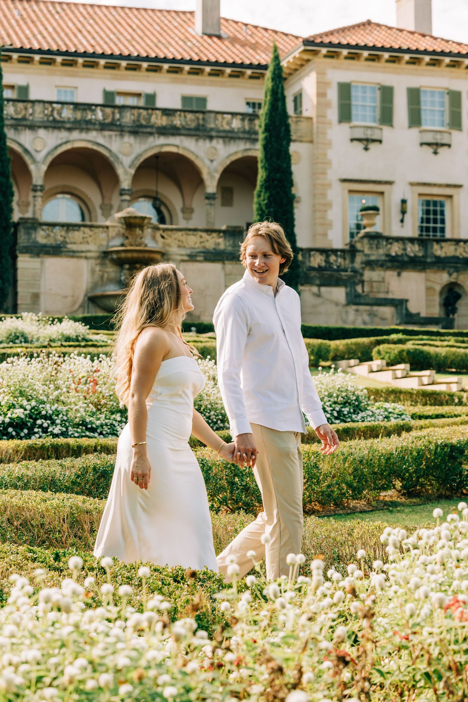 Two people in white outfits walking through a lush garden with Mediterranean architecture and cypress trees in background.