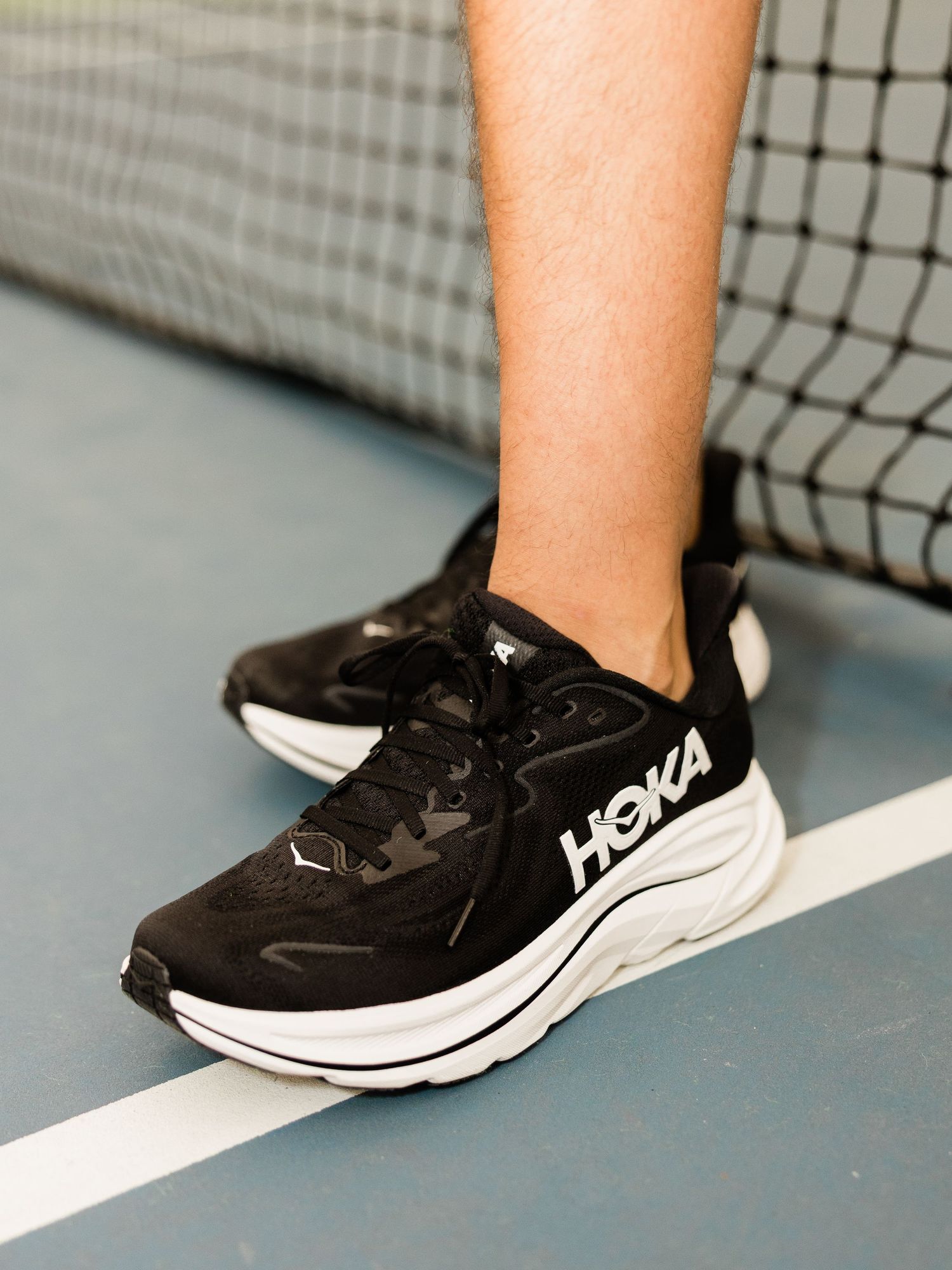 Black and white HOKA running shoes worn on a tennis court near the net.